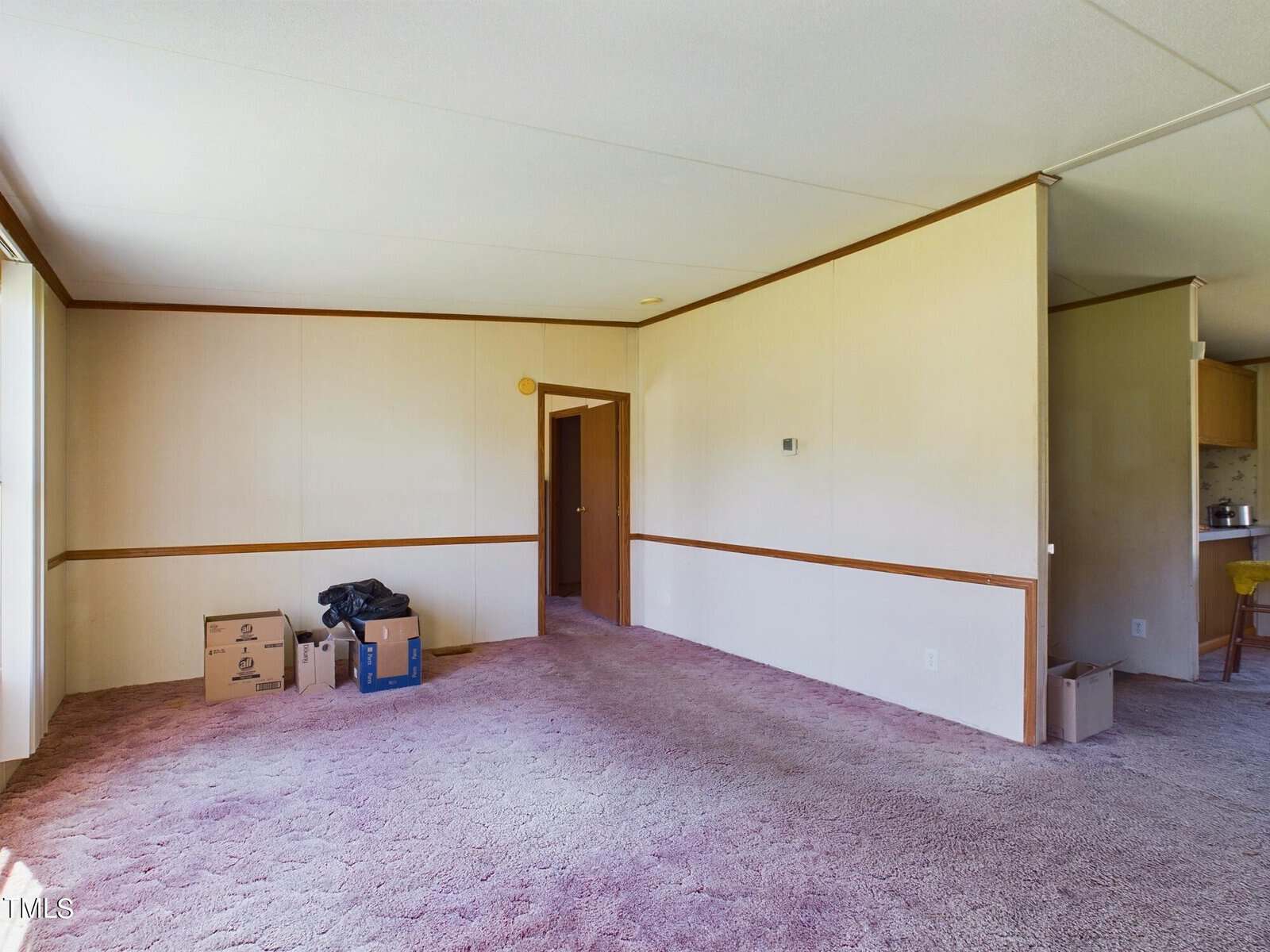 A vacant living room with beige walls, brown trim, and a textured purple carpet. Several cardboard boxes are stacked in a corner. A doorway leads to another room, and part of a kitchen with a countertop is visible in the background.