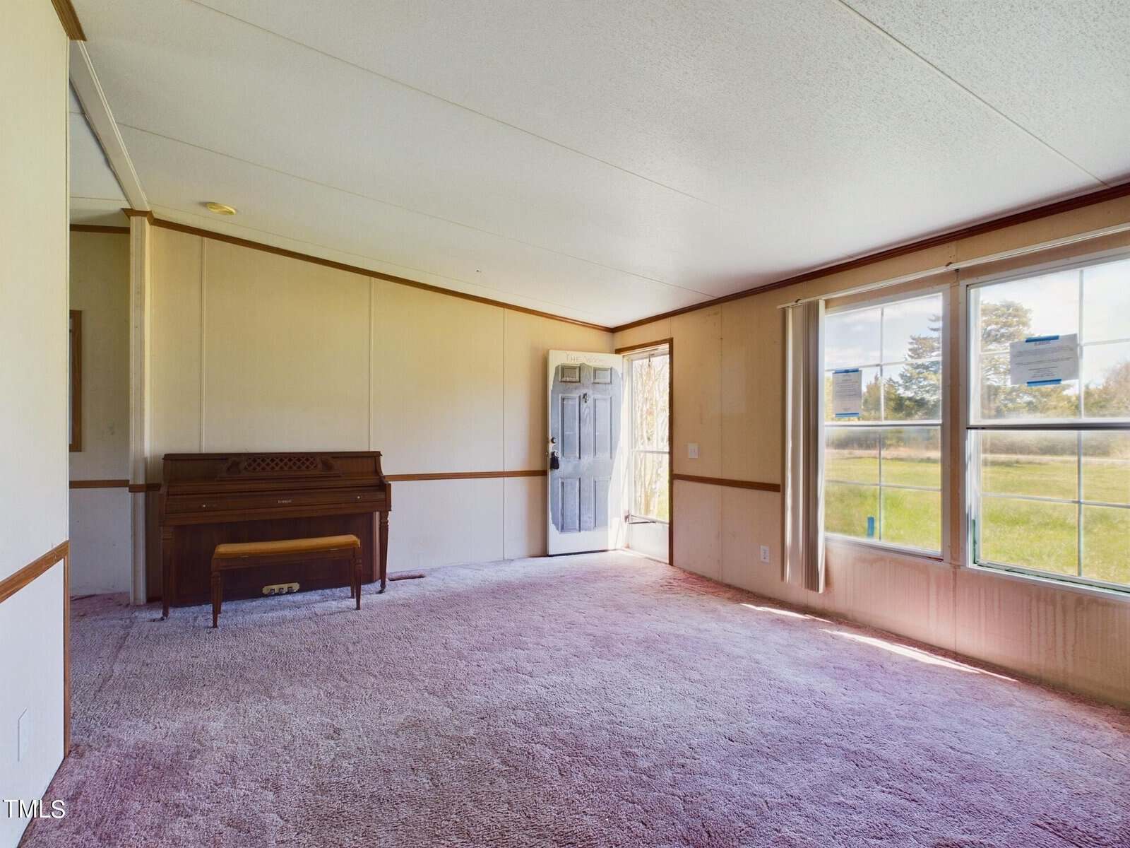 A spacious, unfurnished living room with light-colored walls and carpet. A large window offers views of a lawn and trees outside. A wooden piano sits against one wall, and an open door lets in natural light.