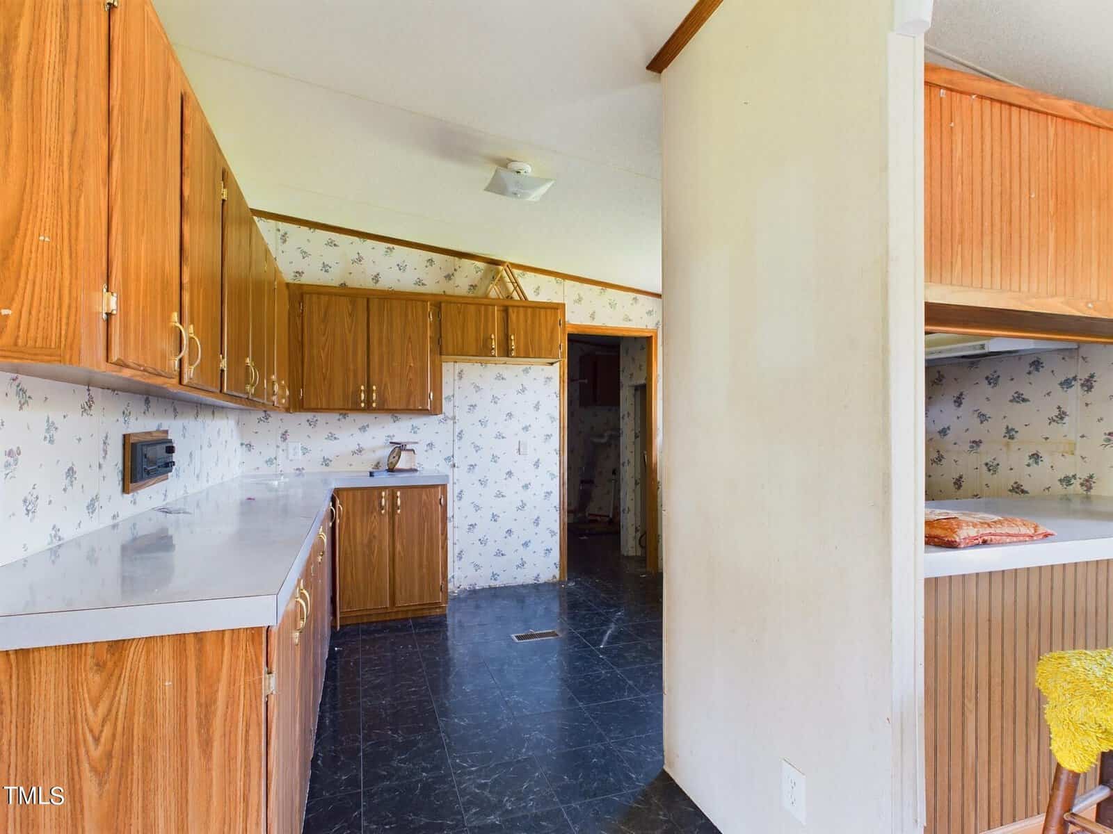A retro-style kitchen with wood cabinets and white countertops. The walls have floral wallpaper, and there's a white fridge. The floor is dark, and a partial view of a breakfast bar is on the right. Lighting is natural and bright.