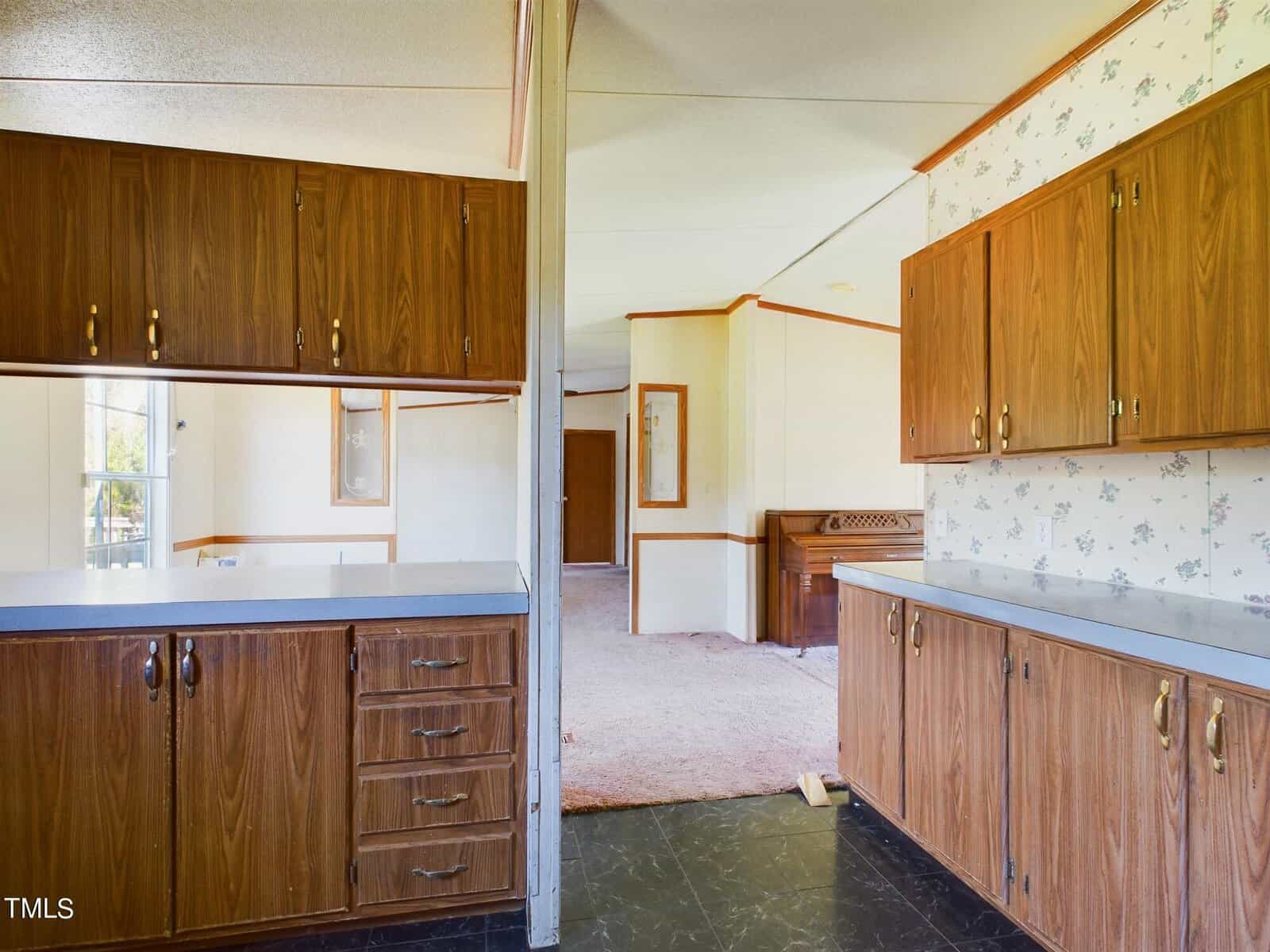 Interior of a home with wood-paneled cabinets on both sides and a floral wallpaper accent in the hallway. The room leads to a carpeted living area, featuring light-colored walls and wood details. Natural light enters from a window in the background.