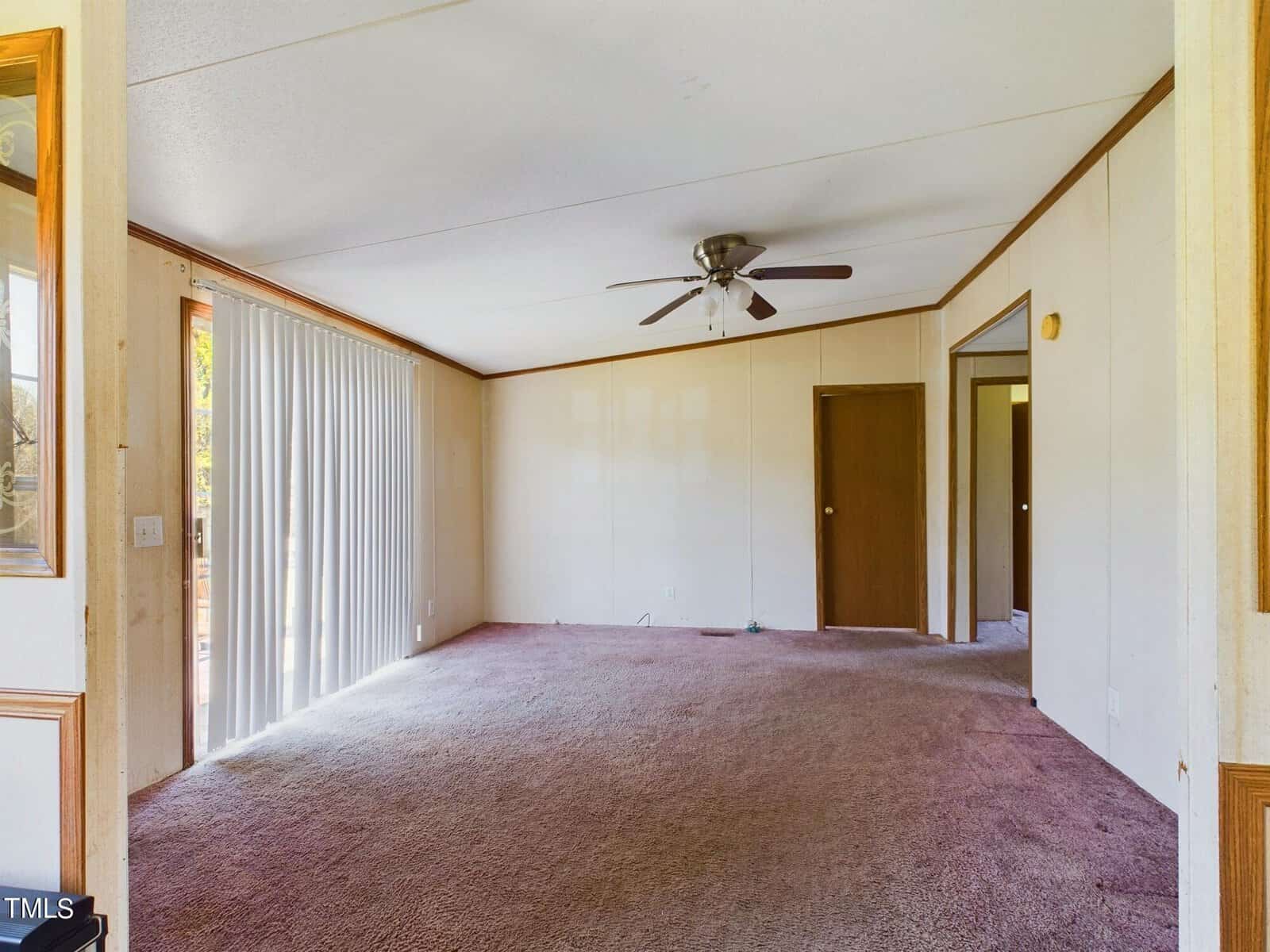 An empty room with beige carpet and walls, featuring a ceiling fan and recessed lighting. Vertical blinds cover the sliding glass doors to the left, and two wooden doors are on the right side. Natural light illuminates the space from the left.