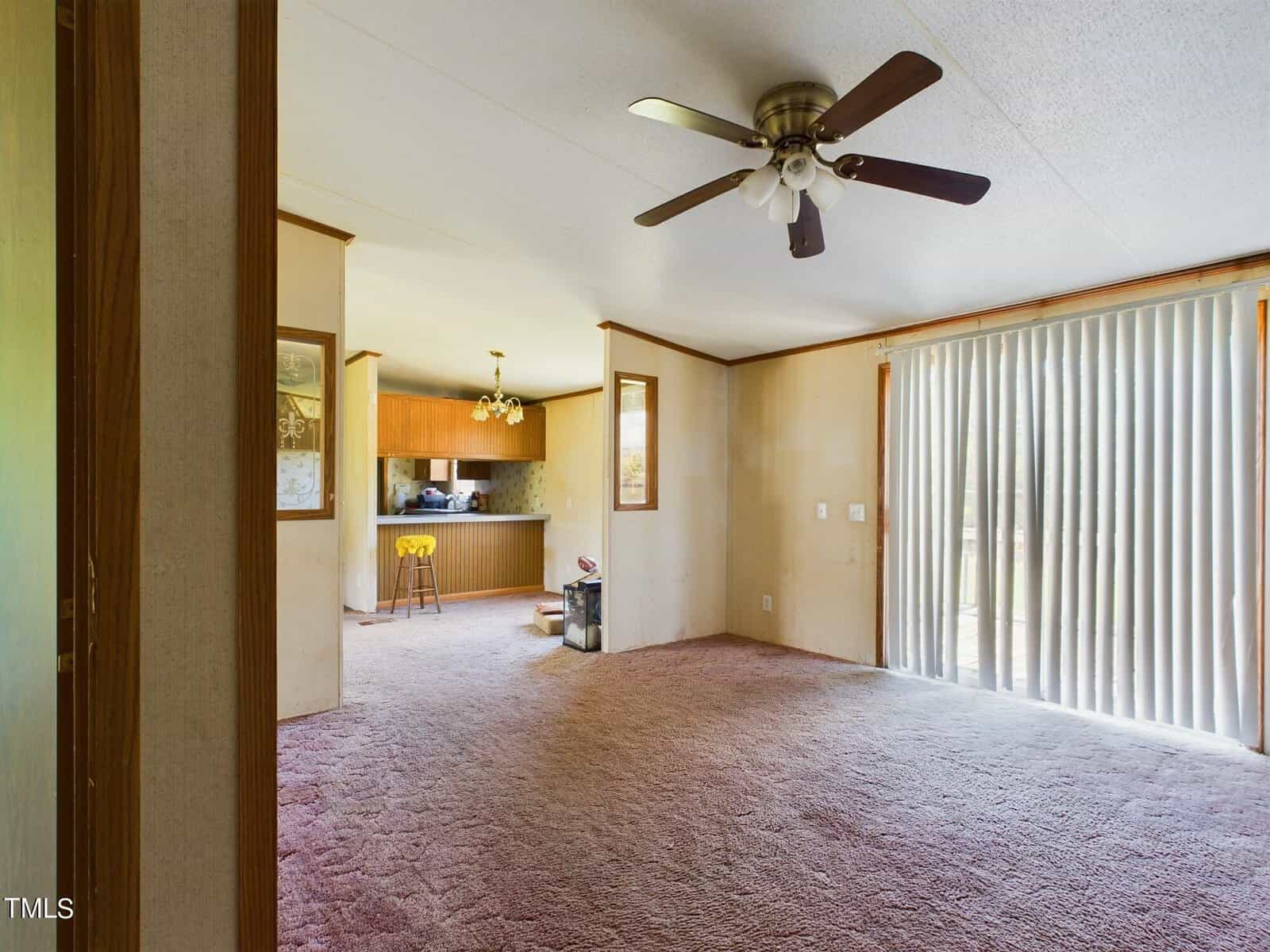 Spacious living room with beige carpet and a ceiling fan. Large sliding glass doors with vertical blinds allow natural light. The room leads to a kitchen area with light wood cabinets and a high stool at the counter.