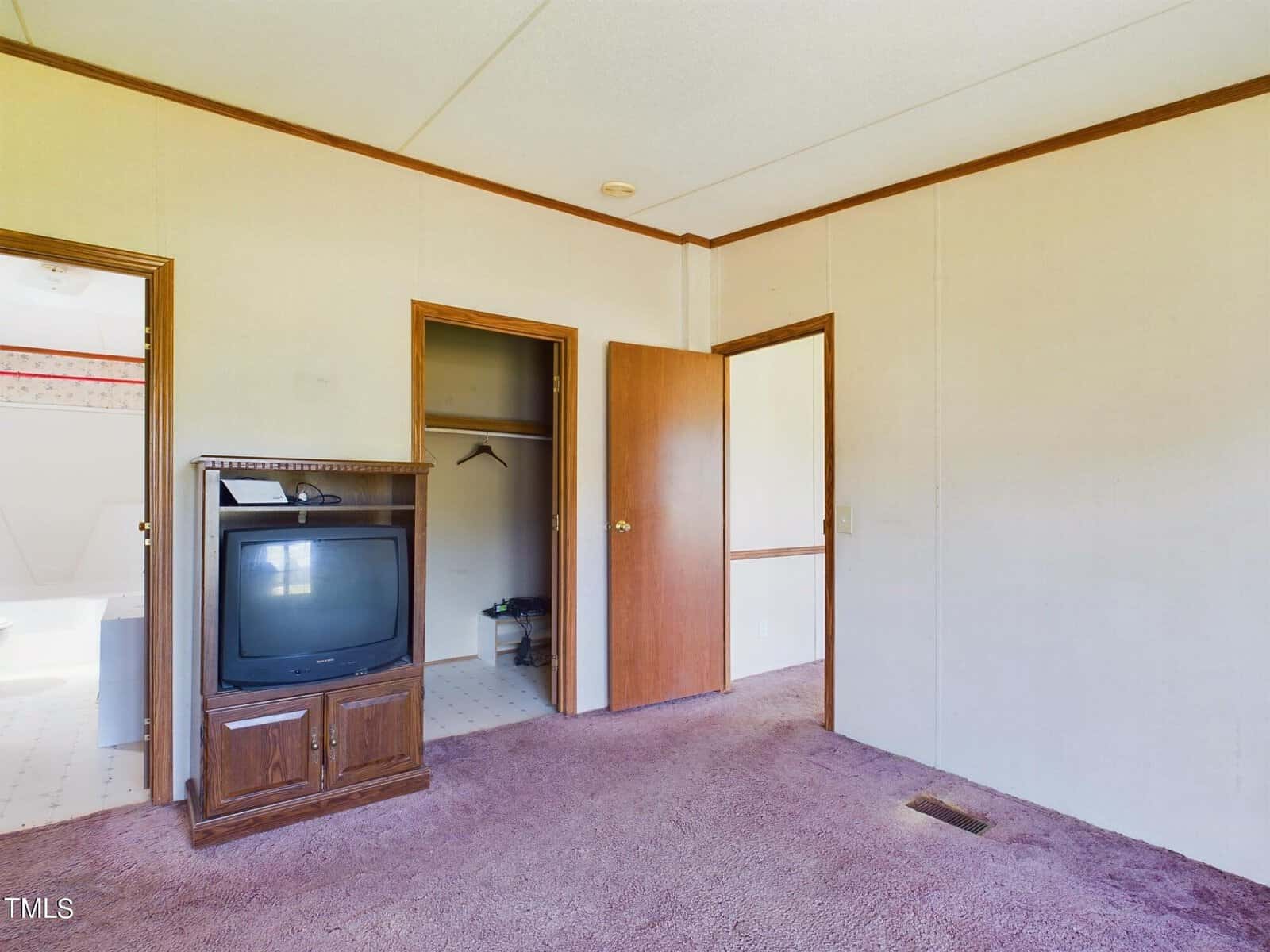 Empty room with beige walls and brown carpet. It has two open doors leading to other areas, a corner shelf with an old TV, and a small closet visible in the doorway. Ceiling is white with a wooden frame around the doors and TV.