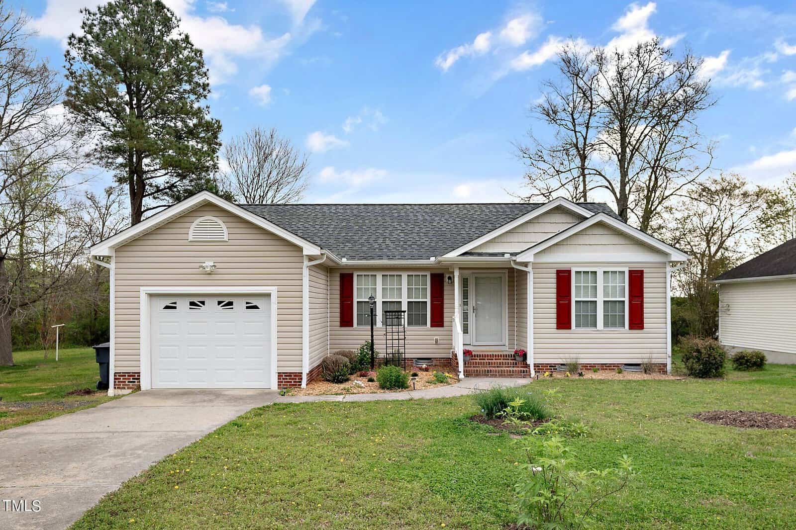 Nestled at 708 Ascot Drive, Creedmoor, this charming single-story house features beige siding, a gray roof, and red shutters. It comes with a one-car garage and a quaint front porch, surrounded by lush grass and a few trees under a partly cloudy sky.
