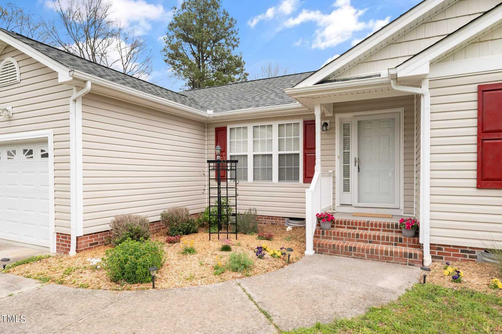 A single-story house with beige siding, a white door, and a small porch. The front yard features a garden with flowers and a bird feeder. A garage is attached on the left. The sky is partly cloudy.
