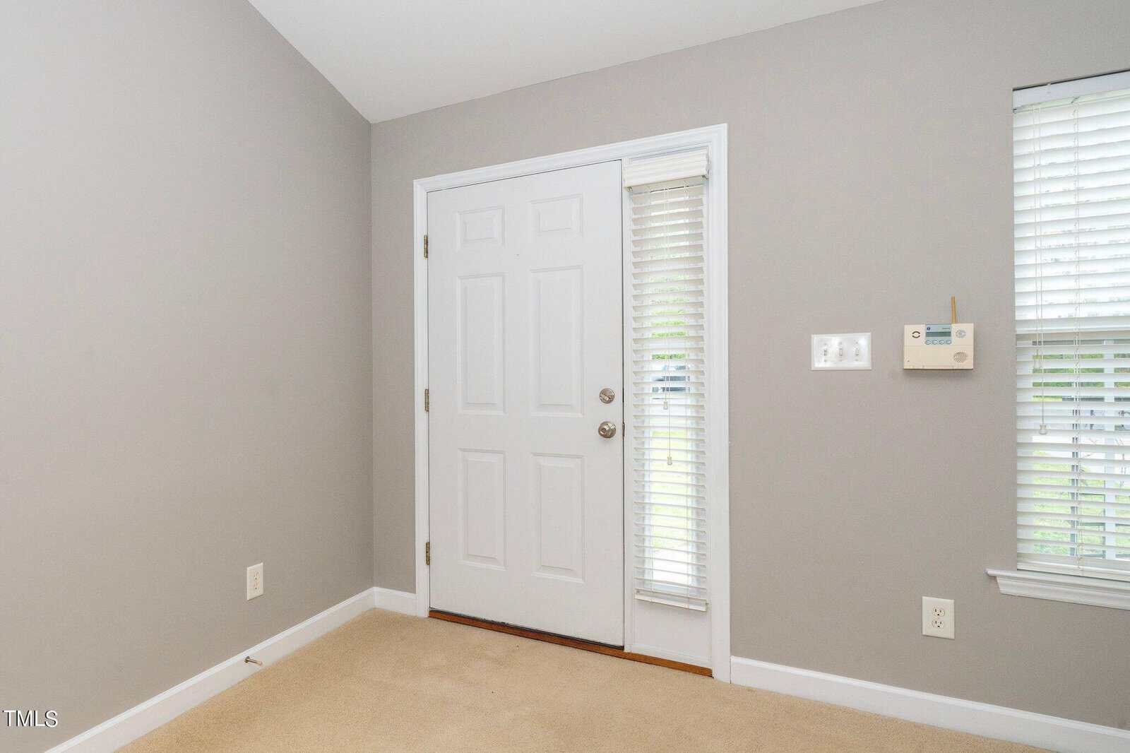 A gray-walled room featuring a white door with an adjacent window covered by white blinds. There's a security keypad on the wall beside the window, and the floor is carpeted in beige.