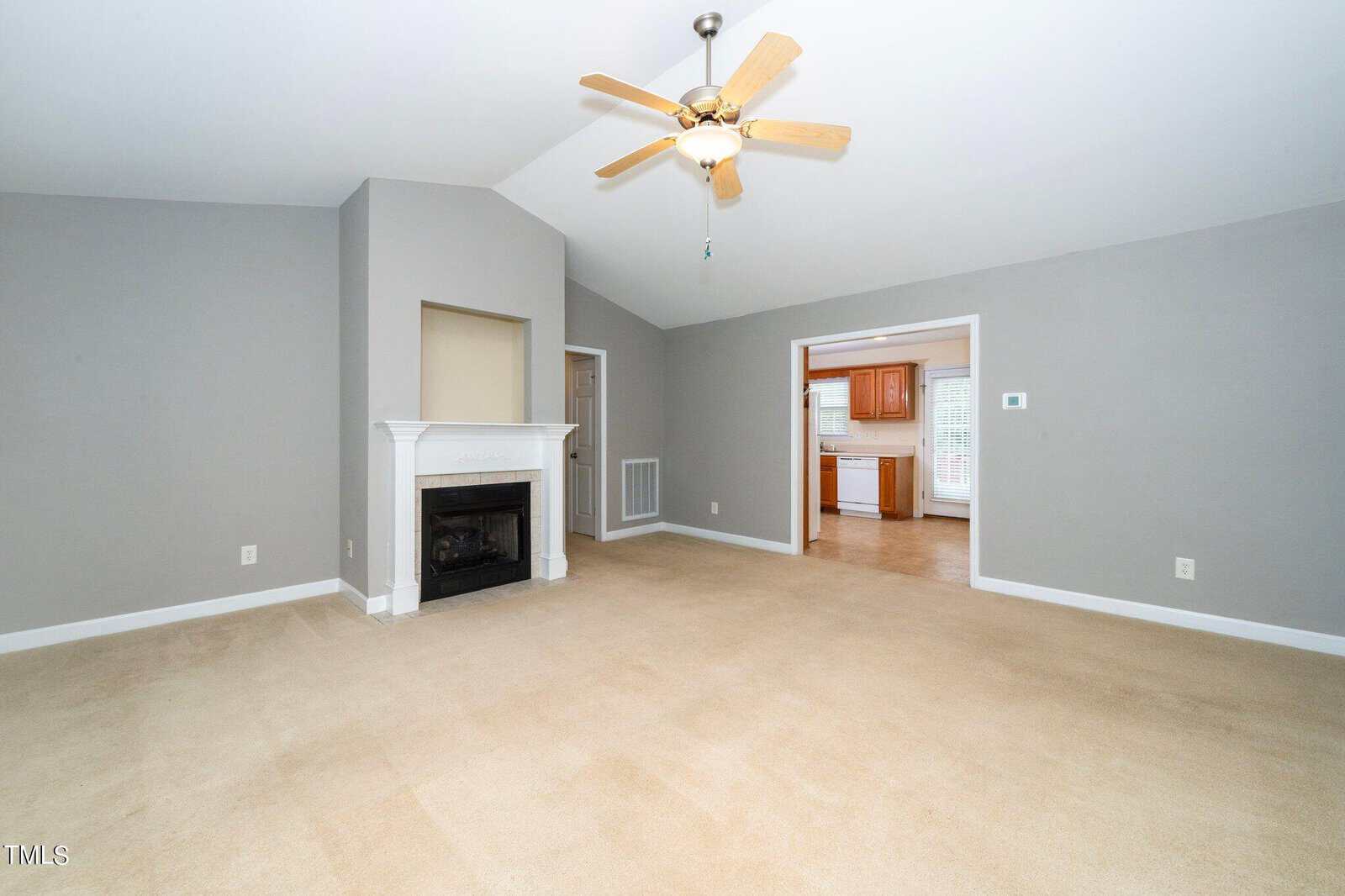 Spacious living room with gray walls and beige carpet. Features a white fireplace with a mantel and a ceiling fan. An open doorway leads to a bright kitchen with wooden cabinets and a window, visible in the background.