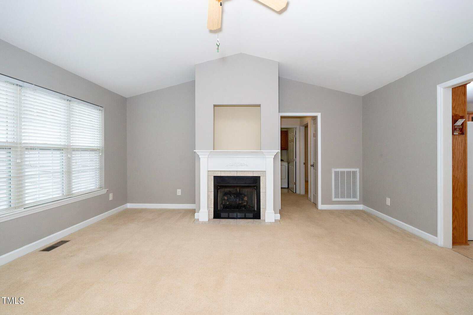 A spacious, empty living room features a vaulted ceiling and a white fireplace centered on a taupe wall. Large windows with blinds provide natural light from the left, and there are doorways on either side of the fireplace. The floor is carpeted in beige.