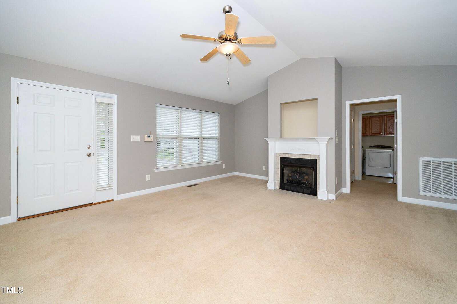 A spacious, unfurnished living room with beige carpet and light gray walls. It features a ceiling fan, a white fireplace, a door leading outside, and an adjacent hallway leading to a laundry area with visible appliances. Natural light comes from a large window.