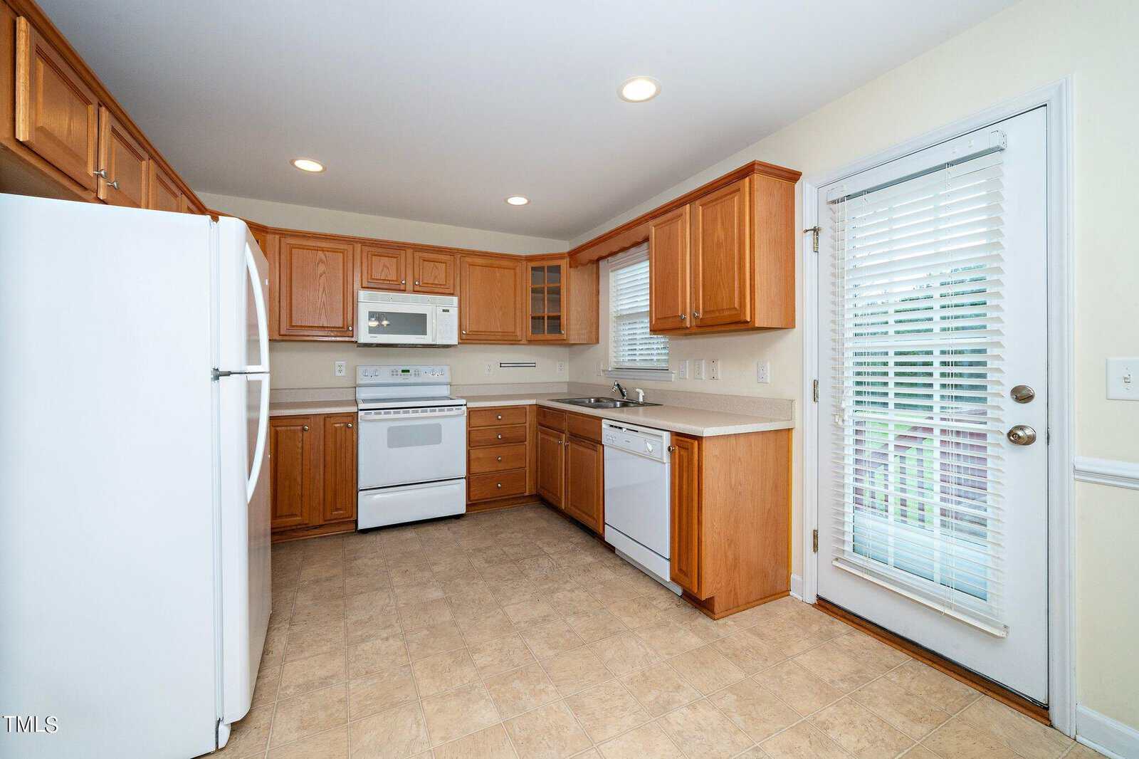 A kitchen with wooden cabinets, a white refrigerator, stove, and dishwasher. It has a beige tile floor, white countertops, and a window with blinds above the sink. A glass door with blinds leads outside, and recessed lighting brightens the room.