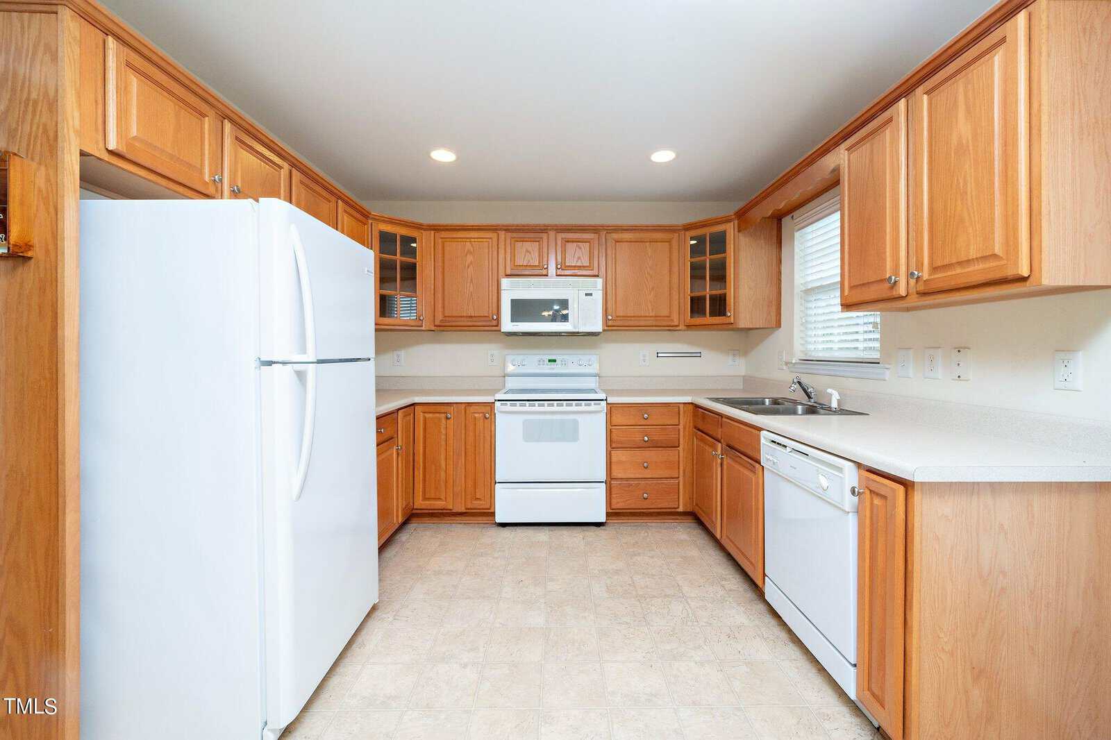 A spacious kitchen with wooden cabinets and white appliances, including a refrigerator, stove, microwave, and dishwasher. The countertops are light-colored, and there are recessed lights in the ceiling. The floor has a beige tile pattern.