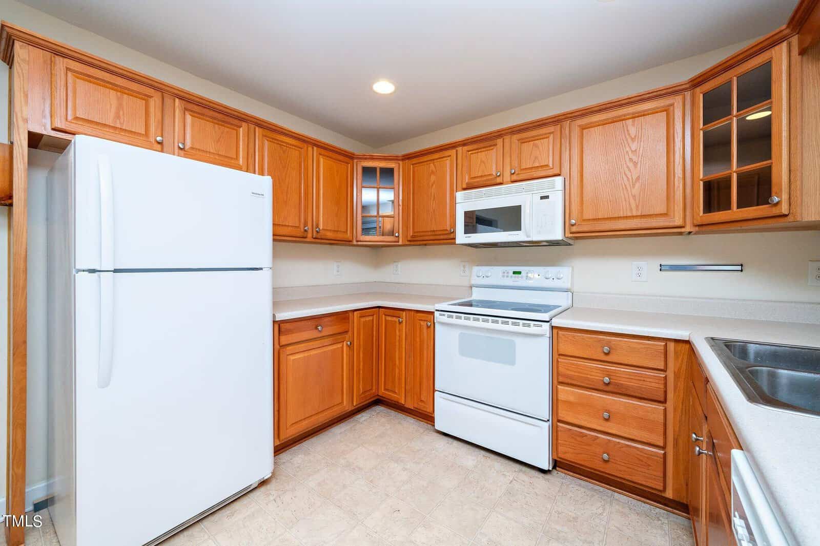A kitchen with wooden cabinets, a white refrigerator, a white electric stove with a microwave above, and a stainless steel sink. The countertops are light-colored, and the walls are neutral, providing a clean and spacious feel.