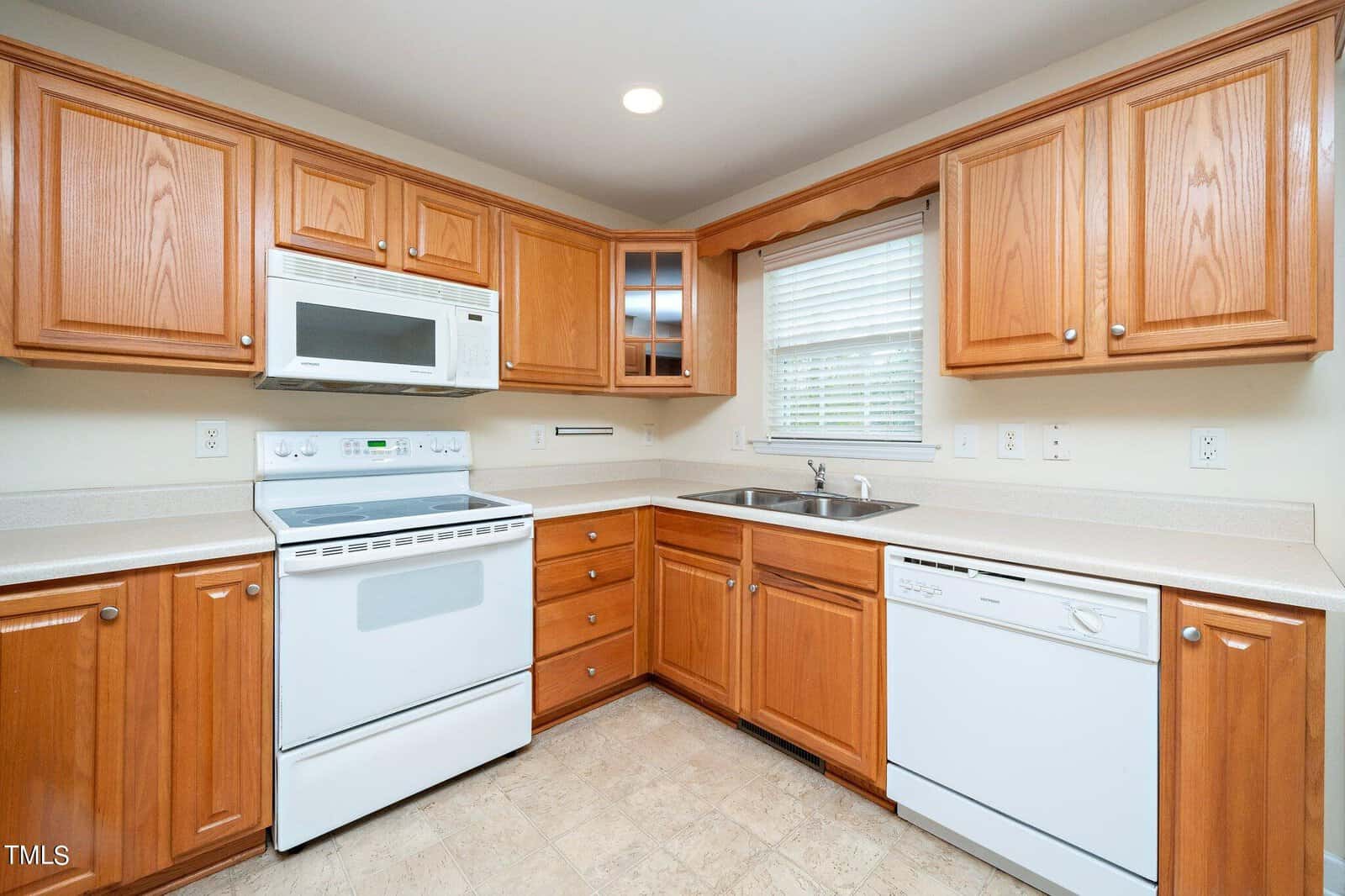 A kitchen with wooden cabinets, an electric stove, a microwave, and a dishwasher. The room features a sink under a window with blinds, light-colored countertops, and tiled flooring.