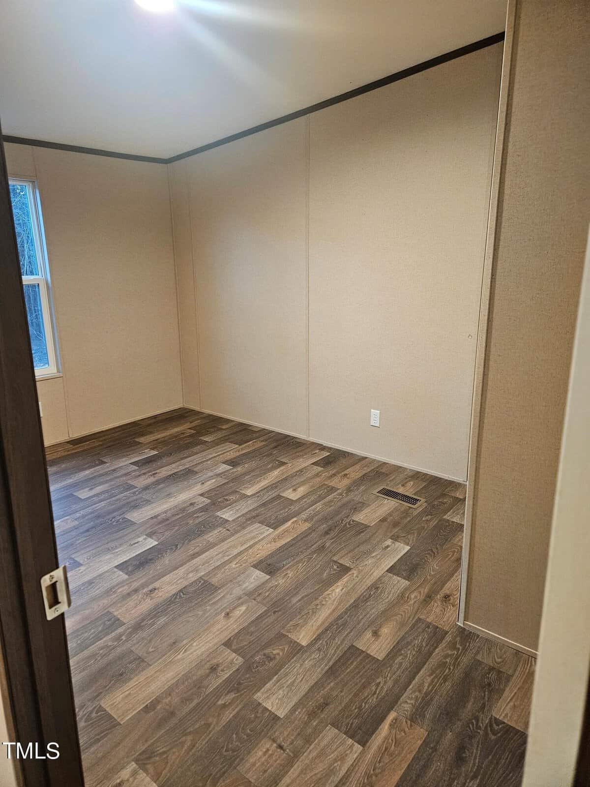 Empty room with beige walls and dark wood-patterned flooring seen from an open door, featuring a large window and minimalistic design.