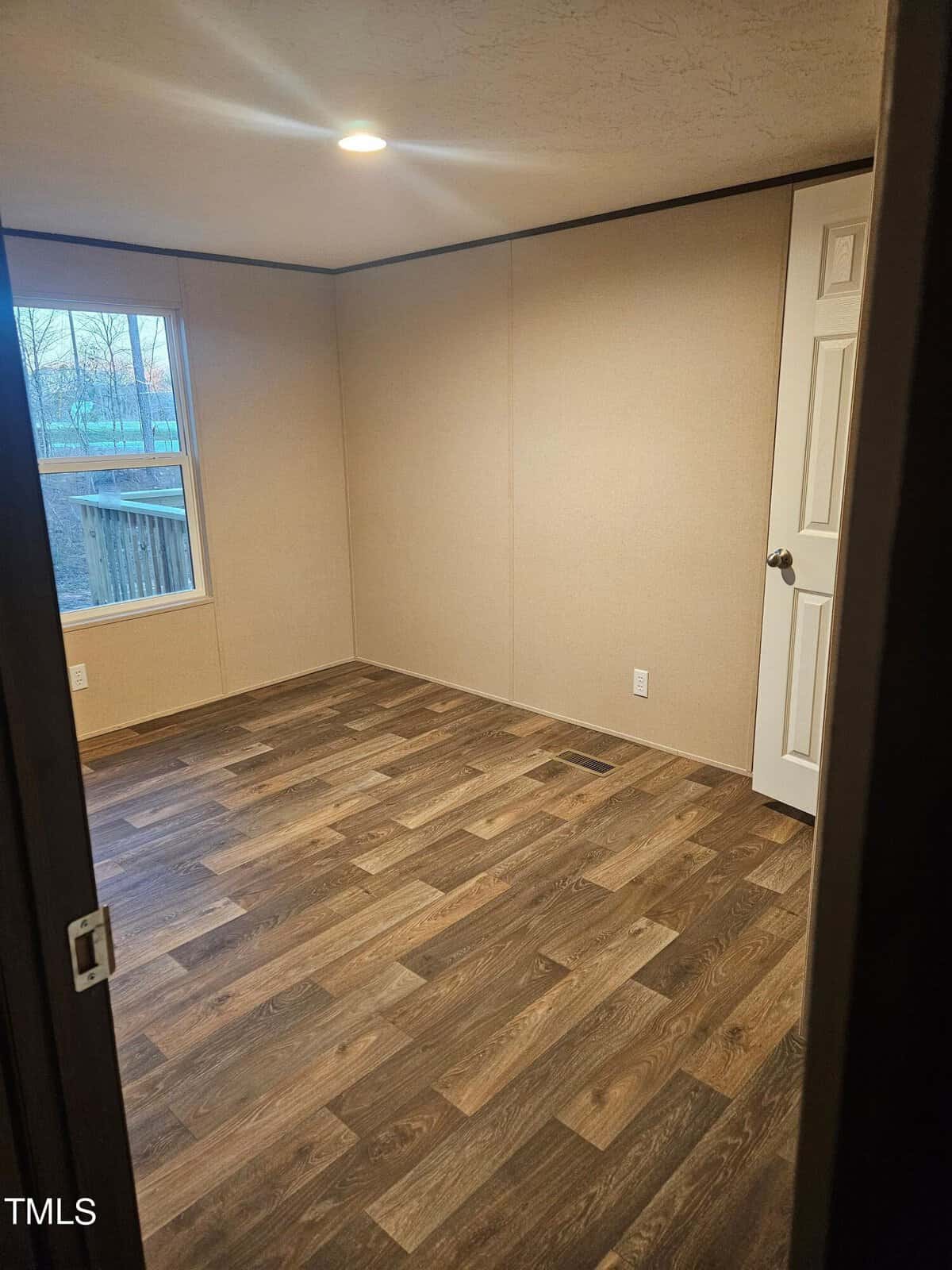 Empty room with light brown walls and dark wood-patterned flooring, featuring a single white door and a window showing outdoor greenery. interior lighting overhead.