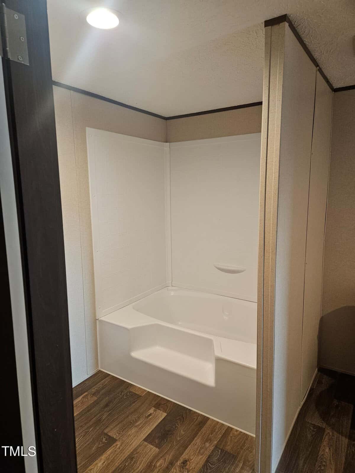 An image of an empty corner shower unit with white walls and a single built-in shelf, installed in a room with wood-effect flooring and partial view of a door frame.