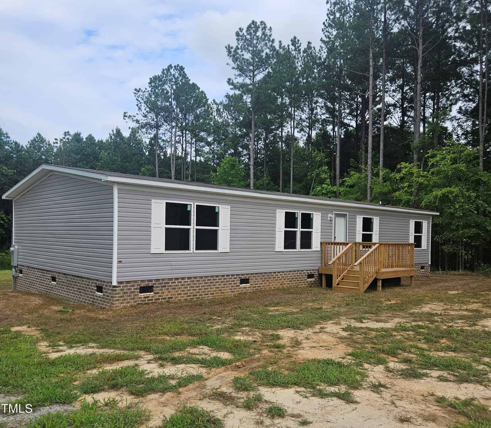 A gray manufactured home with white trim and brick skirting sits on 1212 Flemingtown Road in Henderson, nestled in a clearing surrounded by tall pine trees under a partly cloudy sky. It features a small wooden porch with steps leading to the front door.