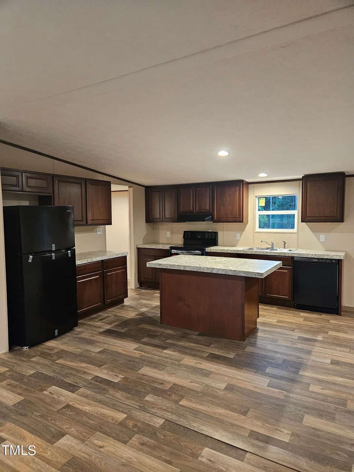 A modern kitchen features dark wooden cabinets, a black refrigerator, a microwave, and an oven. A central island with a granite countertop provides additional workspace. The room has wood-patterned flooring and recessed lighting, and a single window above the sink.