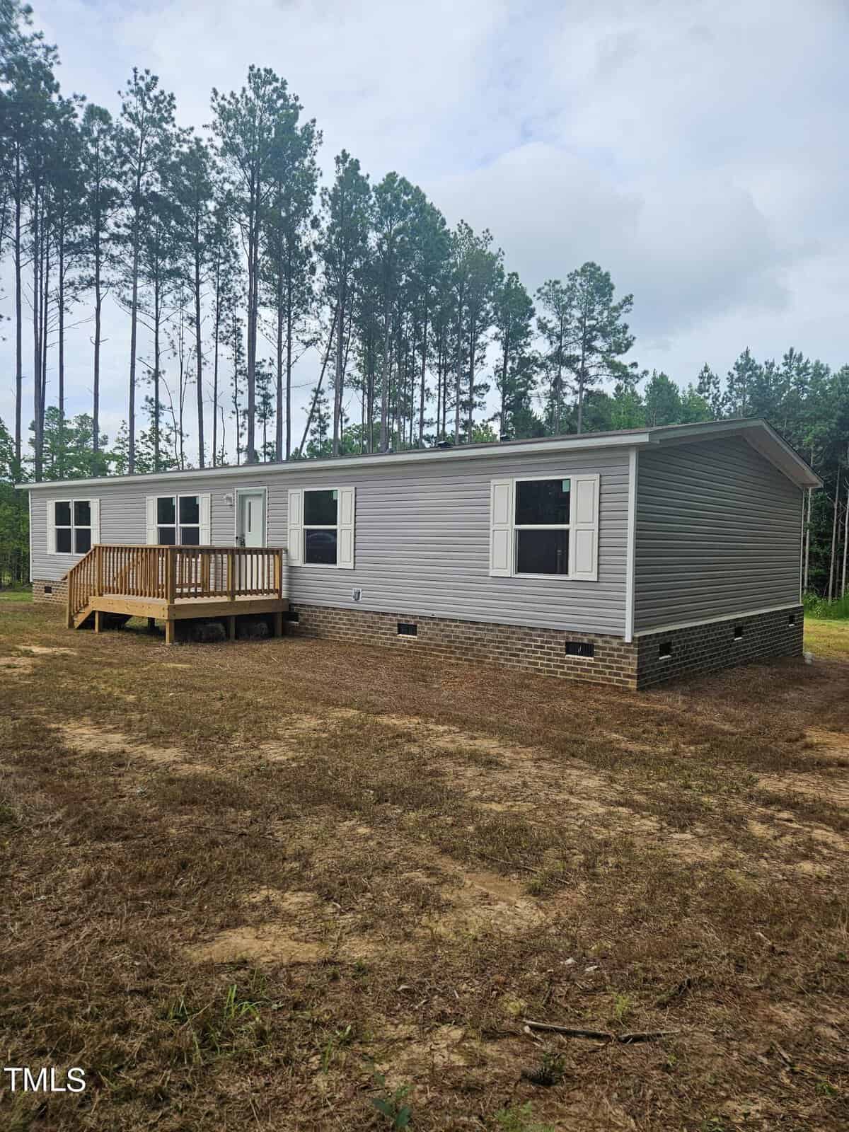 A single-story manufactured home with gray siding and a small wooden porch. It is elevated on a brick foundation and set against a backdrop of tall pine trees, with a patch of bare ground in the foreground.