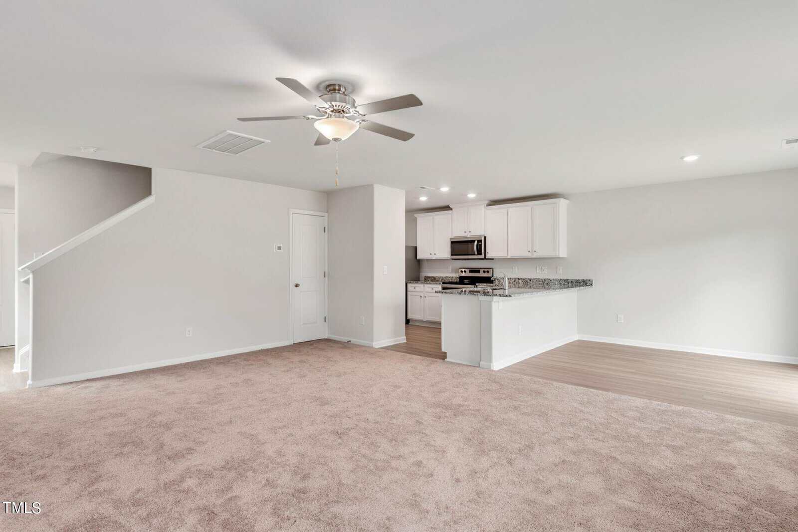 Open-plan living area with beige carpet, a ceiling fan, and neutral walls; adjacent to a modern kitchen with white cabinets, granite countertops, stainless steel appliances, and wood-style flooring. Stairs are visible on the left.