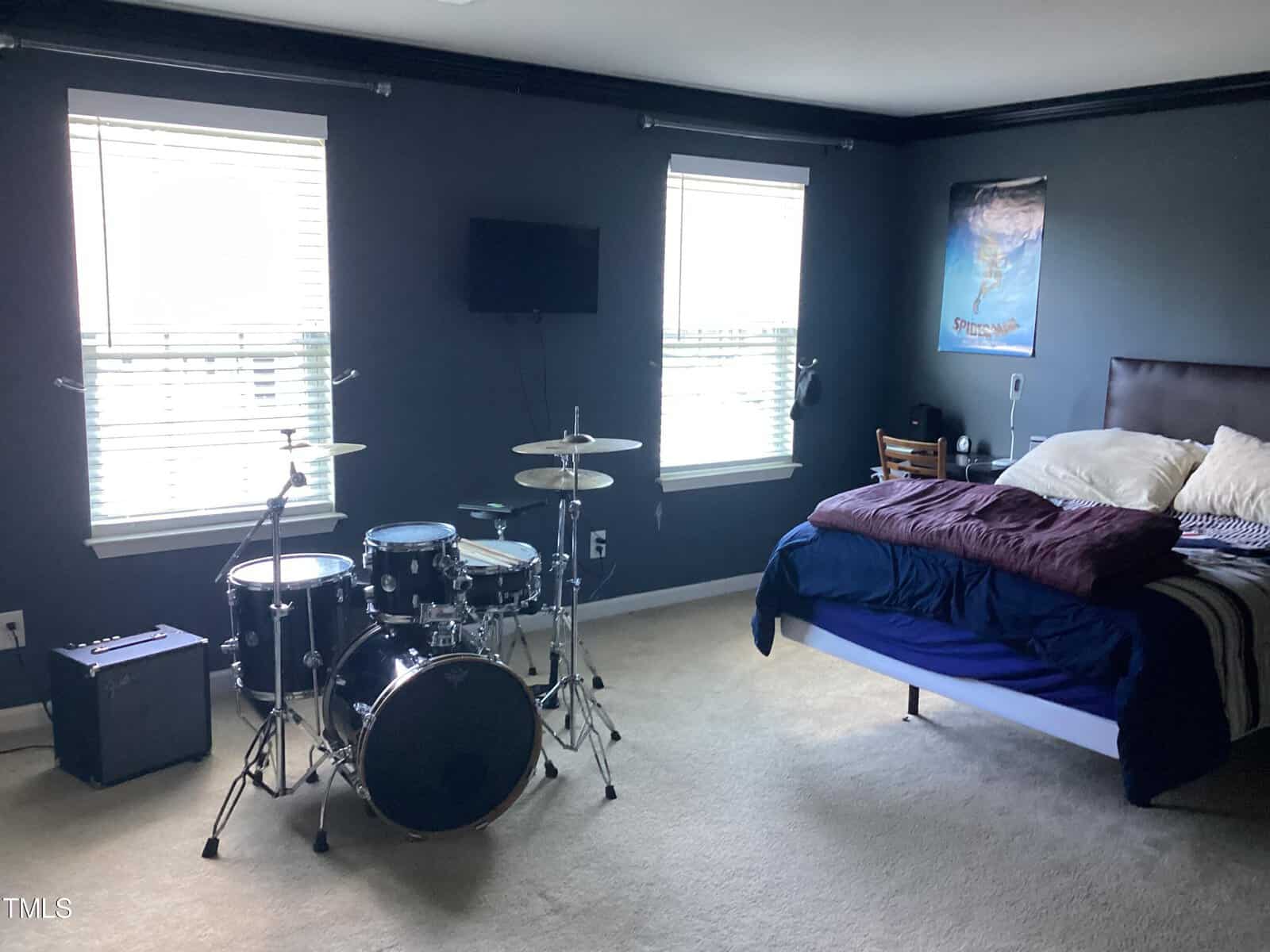 A modern bedroom with dark walls, featuring a neatly made bed with blue bedding and a drum set beside it. a tv is mounted on the wall, and a poster hangs above the bed.