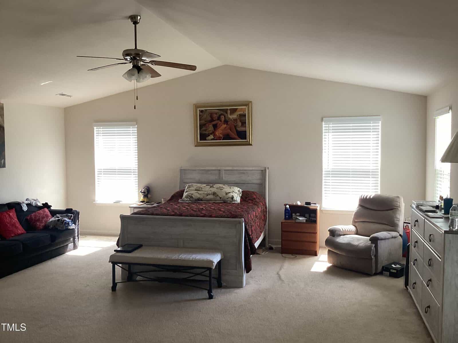 Spacious bedroom with slanted ceiling, large bed with red bedding, two windows with blinds, a ceiling fan, a framed painting above the bed, and a reclining chair near a side table.