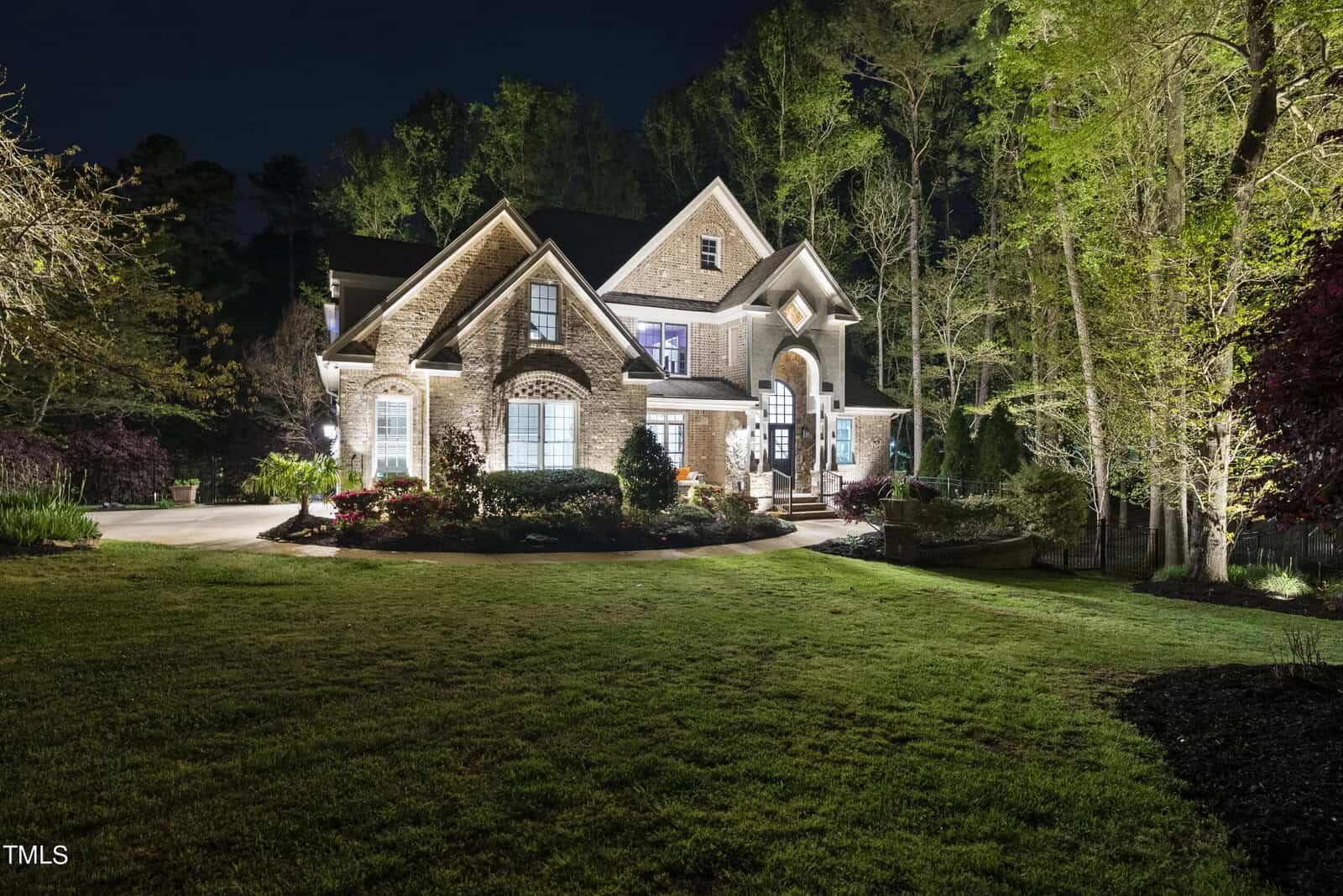 A two-story brick house illuminated at night, surrounded by landscaped greenery and lit trees. The lawn in the foreground is well-maintained, and the house features large windows and a prominent entrance with decorative lighting.