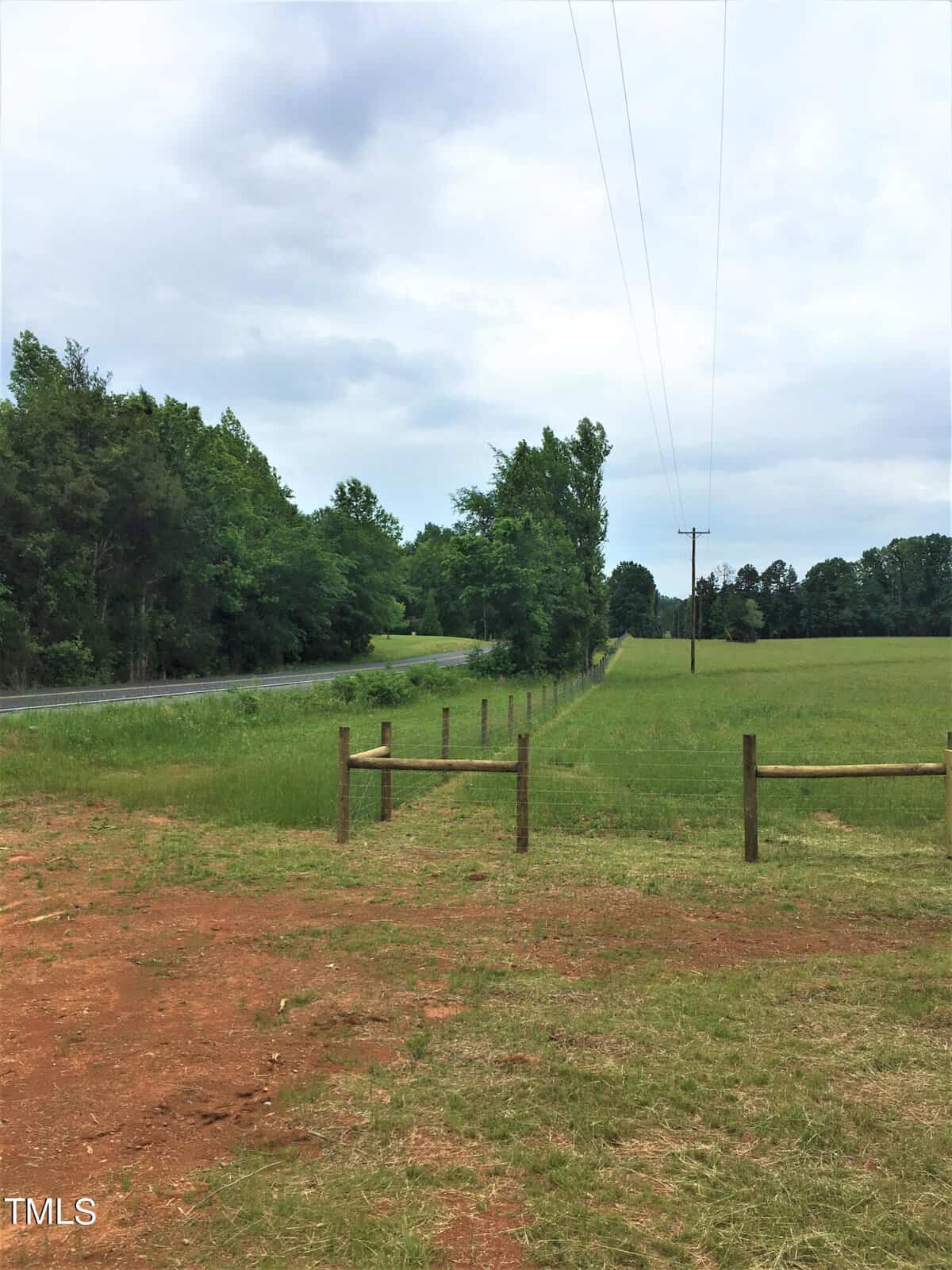 A rural scene with a dirt path, green grassy field, wooden fence, and trees lining a two-lane road under a cloudy sky. Power lines run parallel to the road through the field.