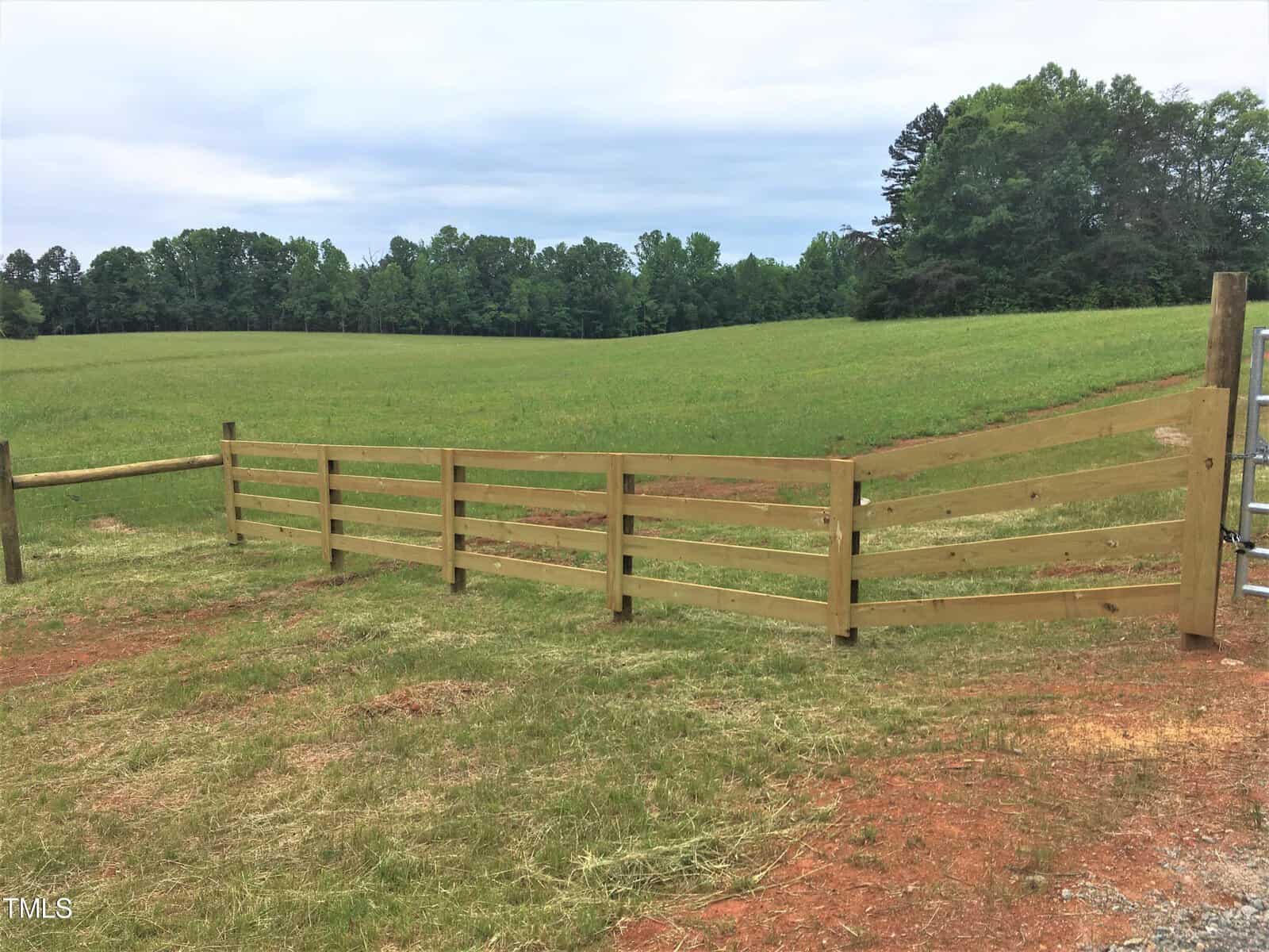 A wooden split-rail fence runs diagonally across a grassy field with a backdrop of trees under a cloudy sky. The ground near the fence is reddish-brown dirt.