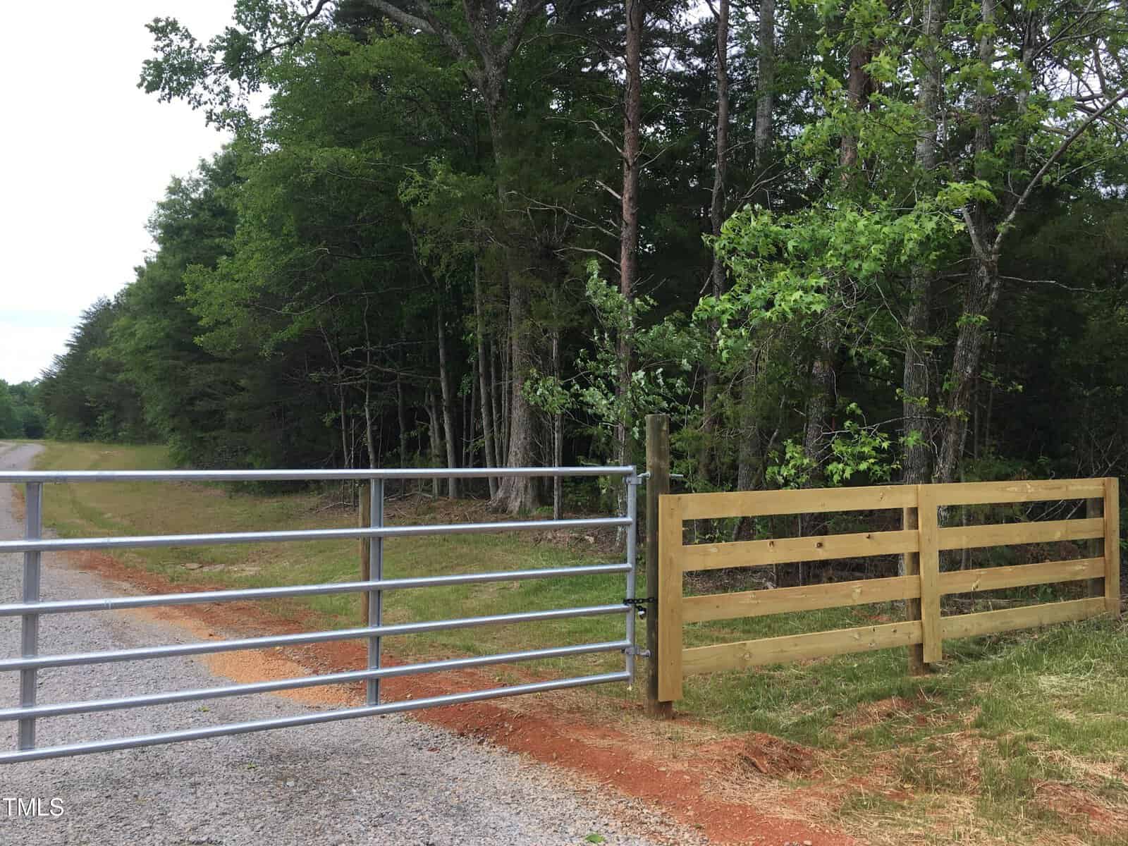A metal gate and wooden fence border a gravel road next to a forested area with dense green trees and grass.