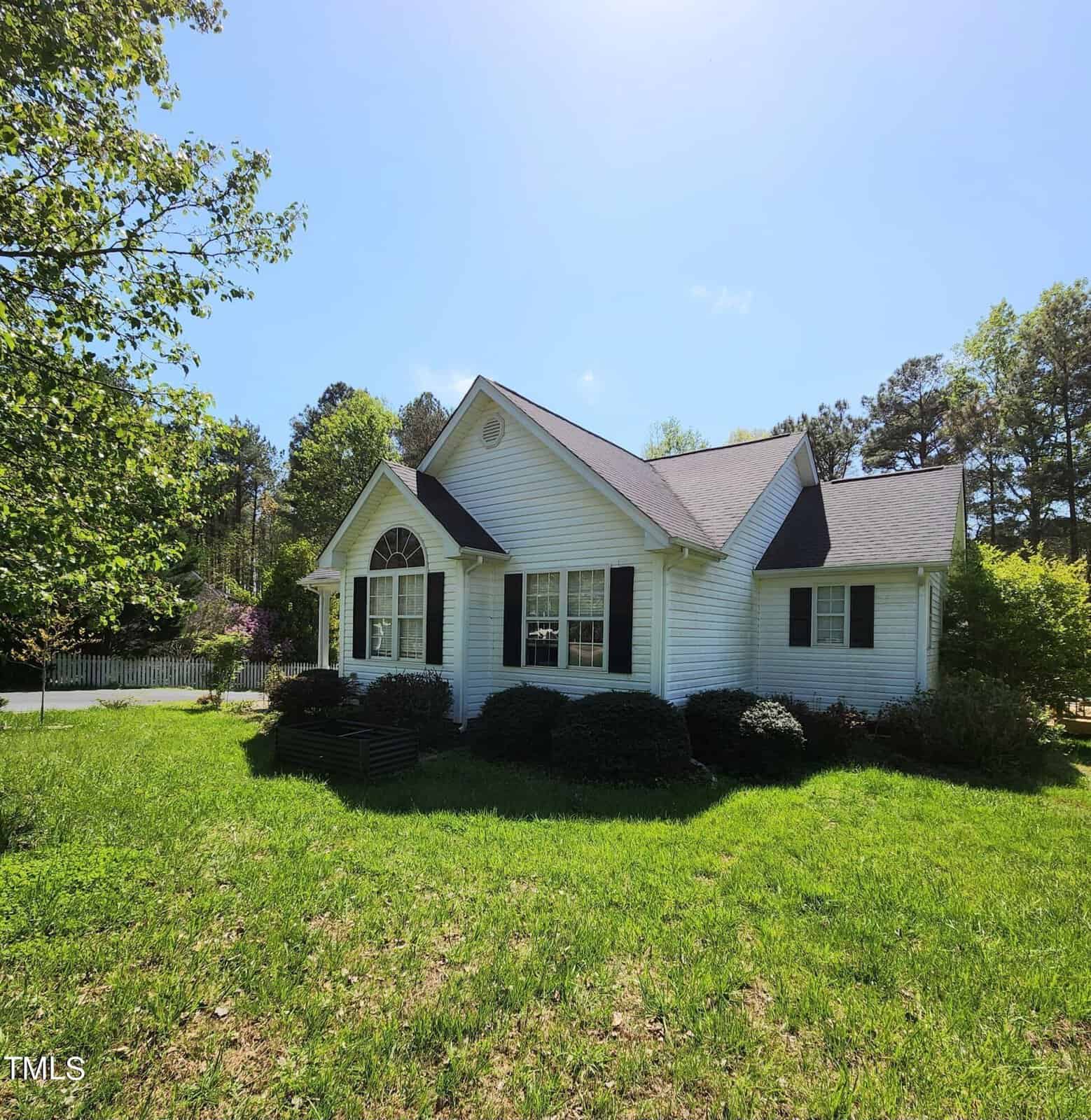 Situated at 3142 Pier Pointe Drive in the charming Creedmoor area, this white single-story house boasts gray shingles and dark shutters. It's surrounded by a lush green lawn and trees, with a large arched window gracing the front. Bushes line the exterior under a clear, sunny sky.