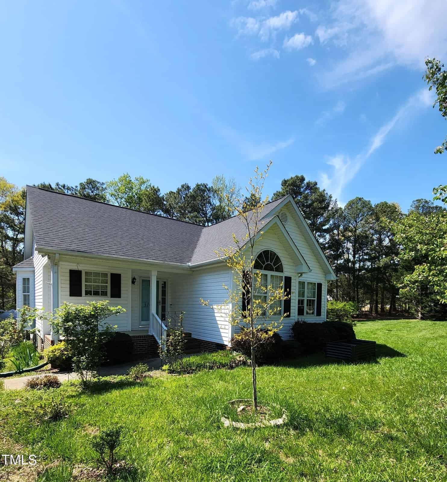 A small white house with a dark roof sits amidst a green lawn, surrounded by trees. The sky is blue with a few clouds, and there is a small, leafless tree in the foreground.