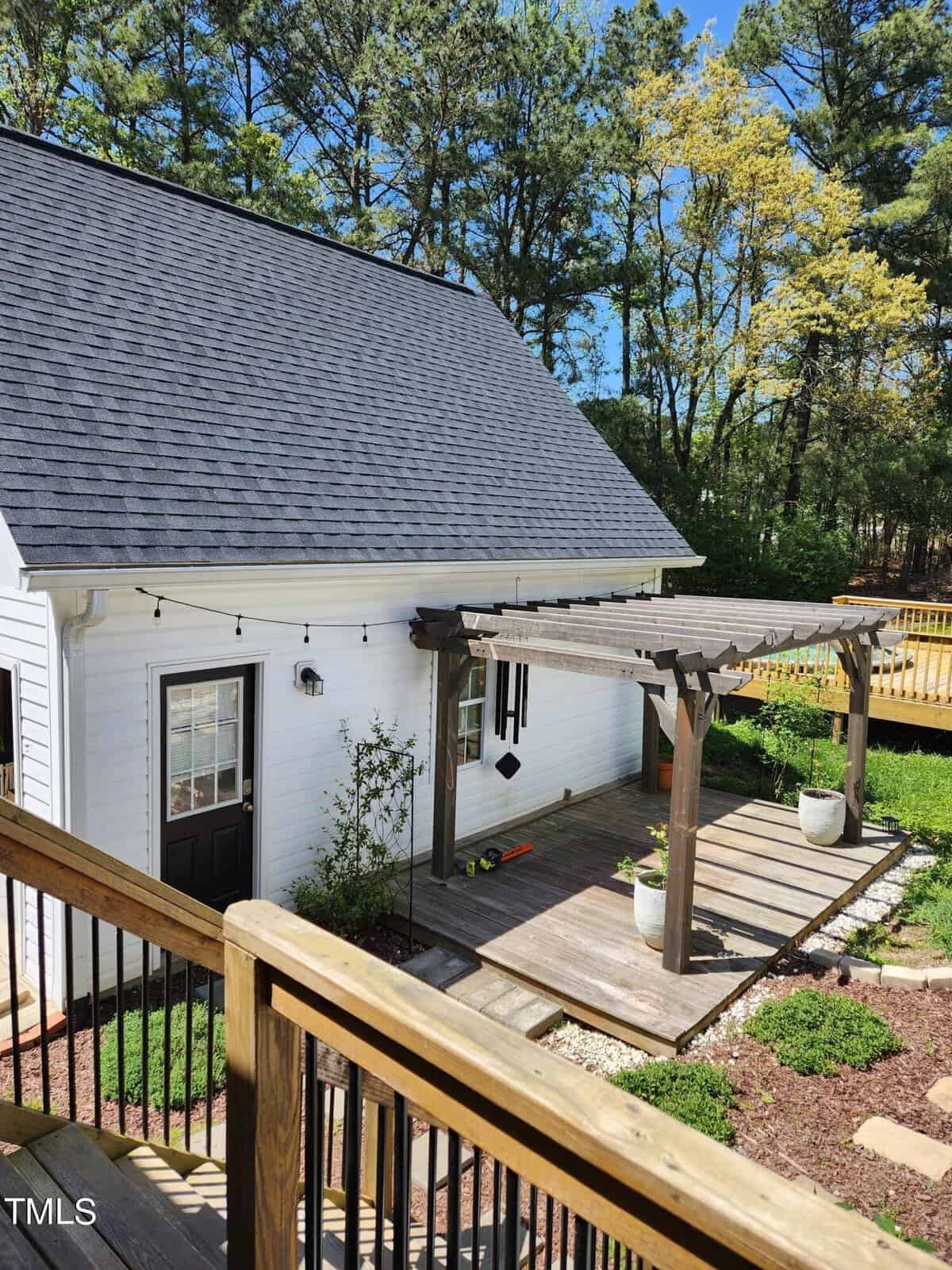 A white house with a dark sloped roof and an attached wooden pergola over a deck. The deck features two large planters. There is a garden area with shrubs and a staircase leading down from the house. Trees and a fence are visible in the background.