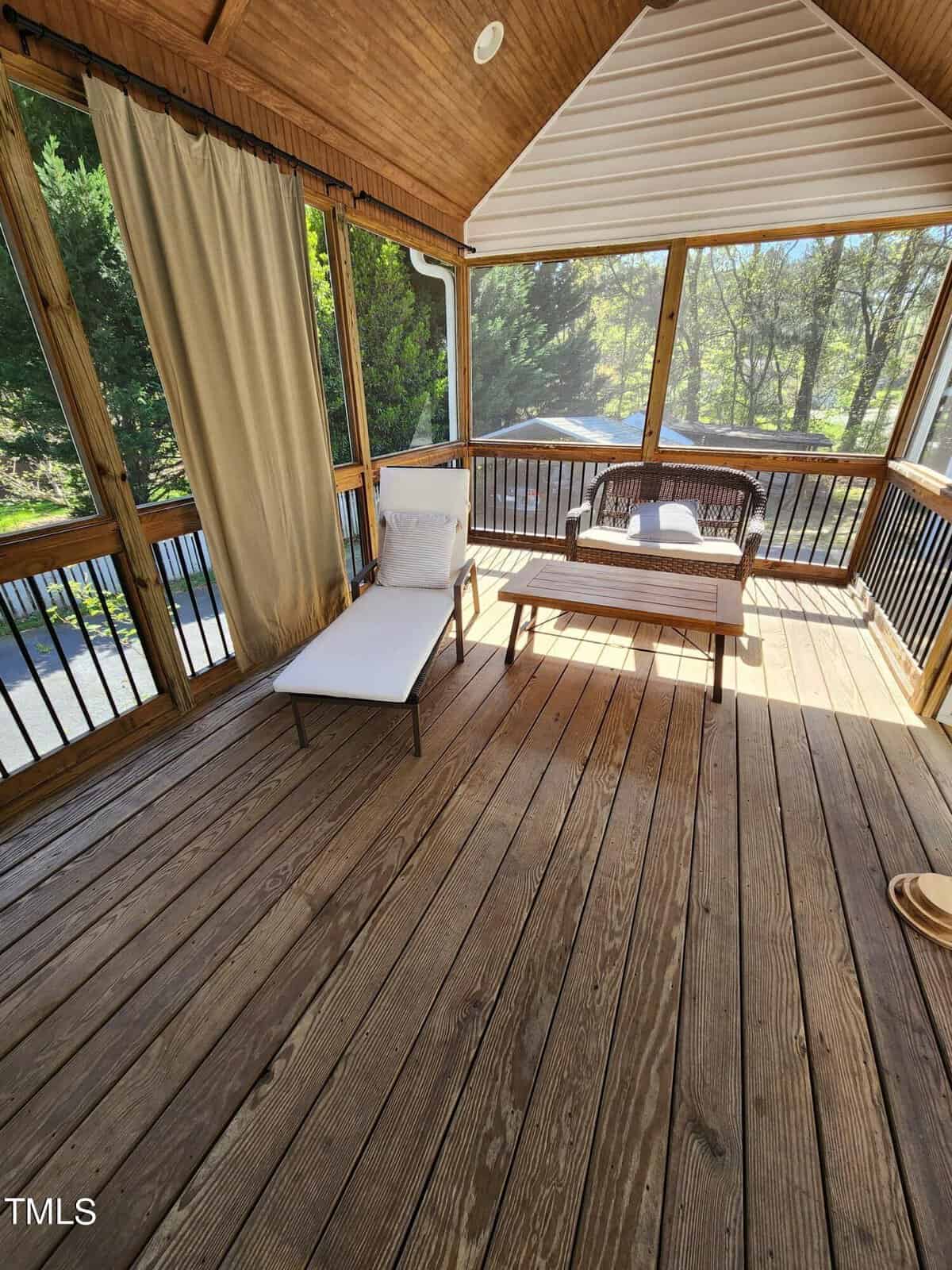 A cozy screened porch with wooden flooring features a white lounge chair and a wooden coffee table. Sunlight filters through the trees, creating a warm, inviting atmosphere.