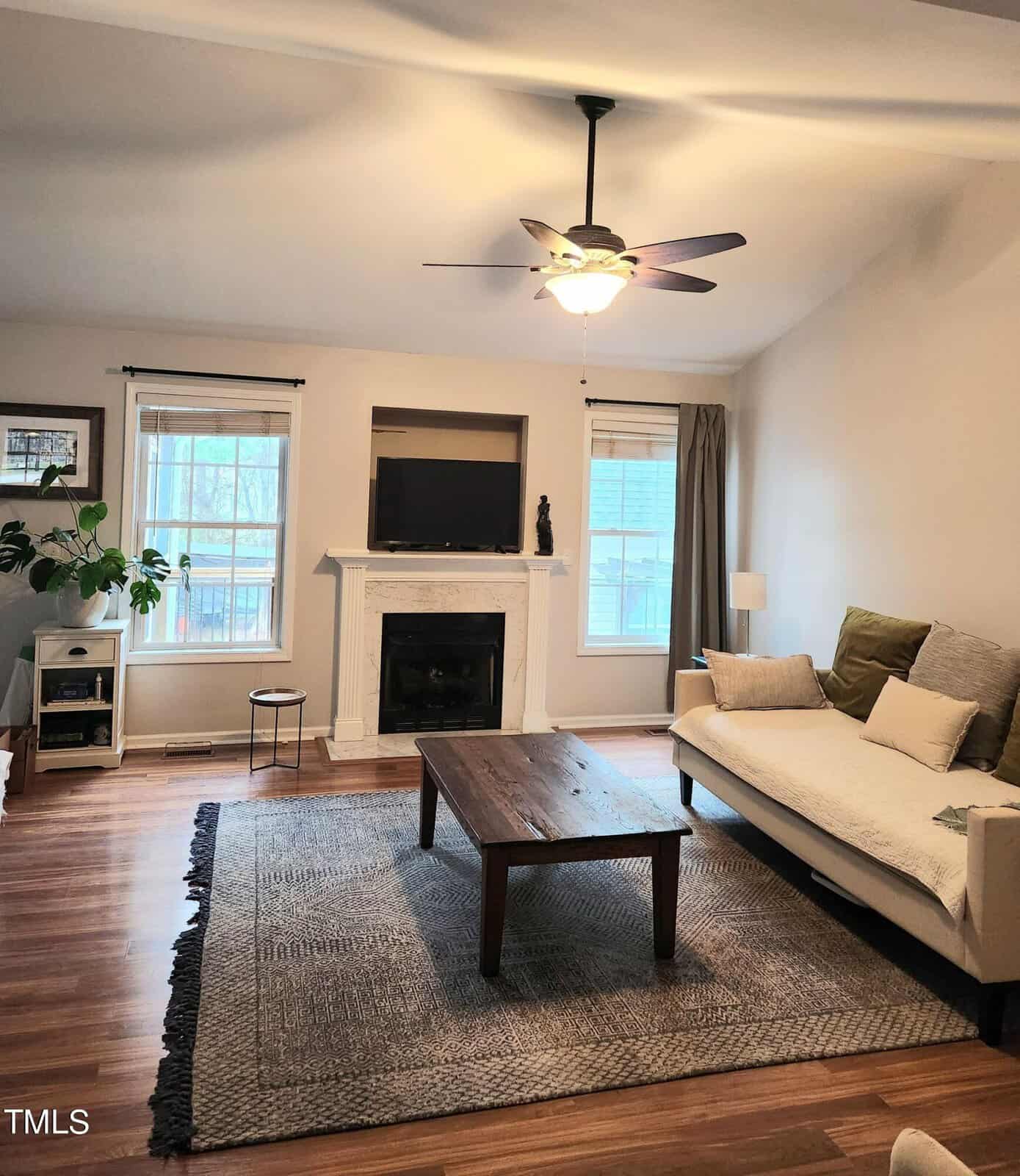 A cozy living room with a ceiling fan, white walls, and a wooden floor. It features a fireplace with a TV on the mantel, a rug, a sofa with green and beige cushions, and a wooden coffee table. A plant and windows complete the scene.