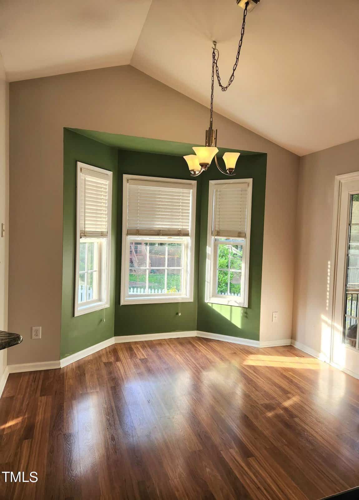A sunlit room with a vaulted ceiling features a bay window with green accents and white blinds. Light wood flooring complements the soft beige walls. A chandelier hangs from the ceiling. A glass door leads to an outdoor area with a fence visible.