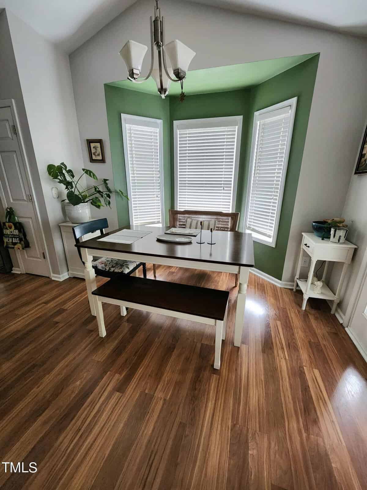 Dining area with a wooden table and bench, placed in front of a bay window with white blinds. Green walls accentuate the space, and a chandelier hangs above. A small white side table and potted plant are on the left. Wood floor throughout.