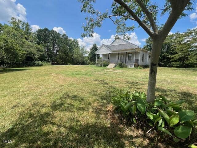 A small white house with a front porch is situated in a spacious grassy yard. A solitary tree with some plants at its base stands in the foreground, while a wooded area is visible in the background under a partly cloudy sky.