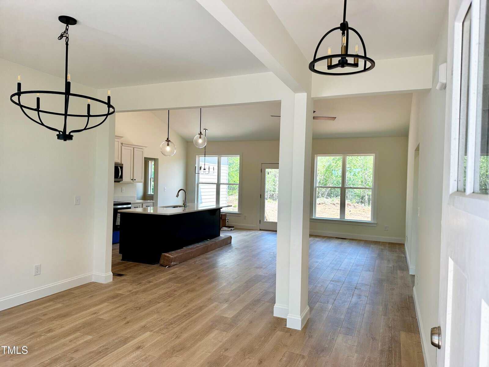 Open-concept living and dining area with wood flooring, white walls, modern black chandeliers, and large windows letting in natural light; kitchen with island visible in background.