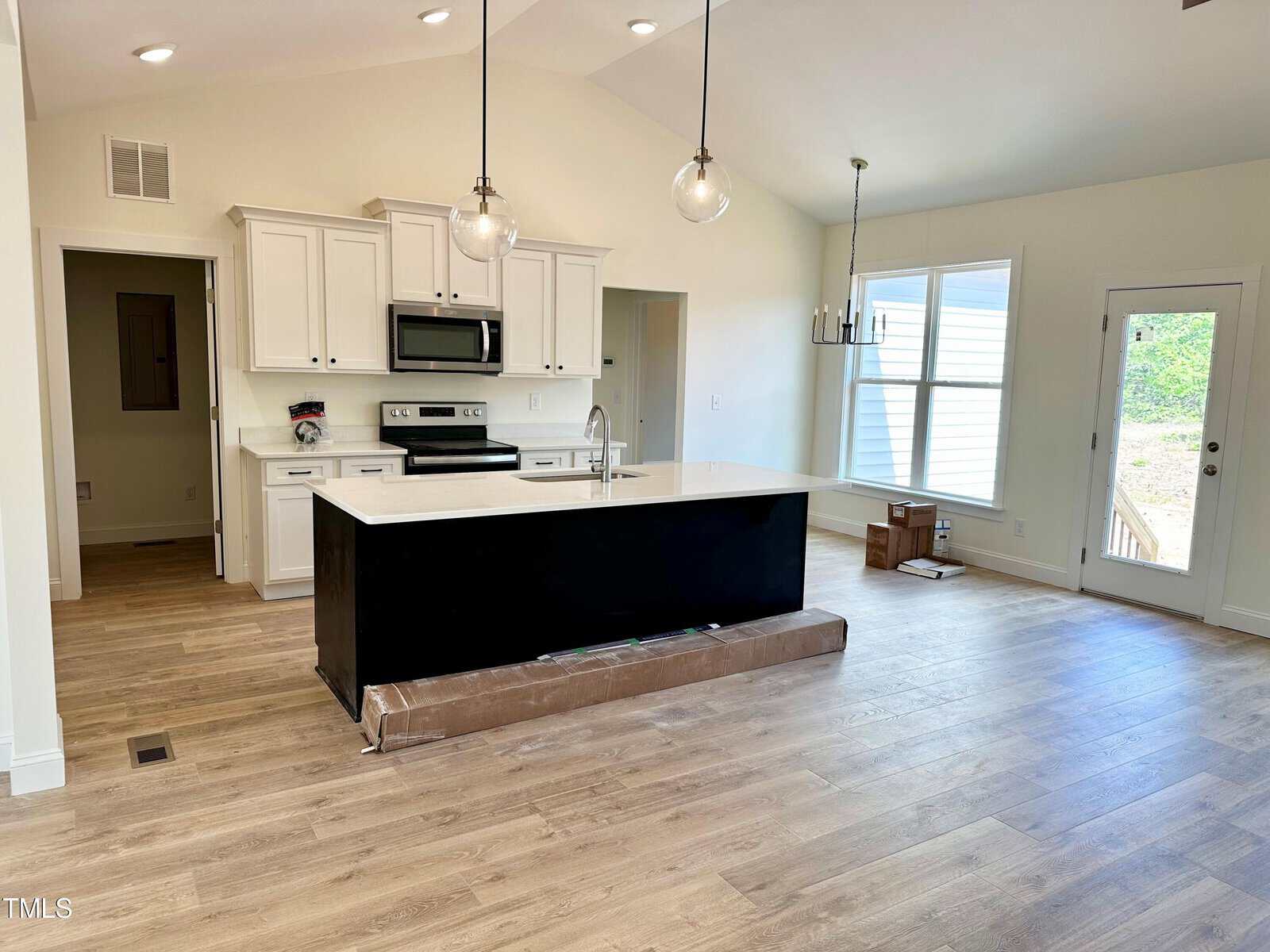 Modern kitchen with white cabinets, stainless steel appliances, a central island with a black base, pendant lights, wood flooring, large windows, and some unpacked moving boxes on the floor in a spacious open layout.