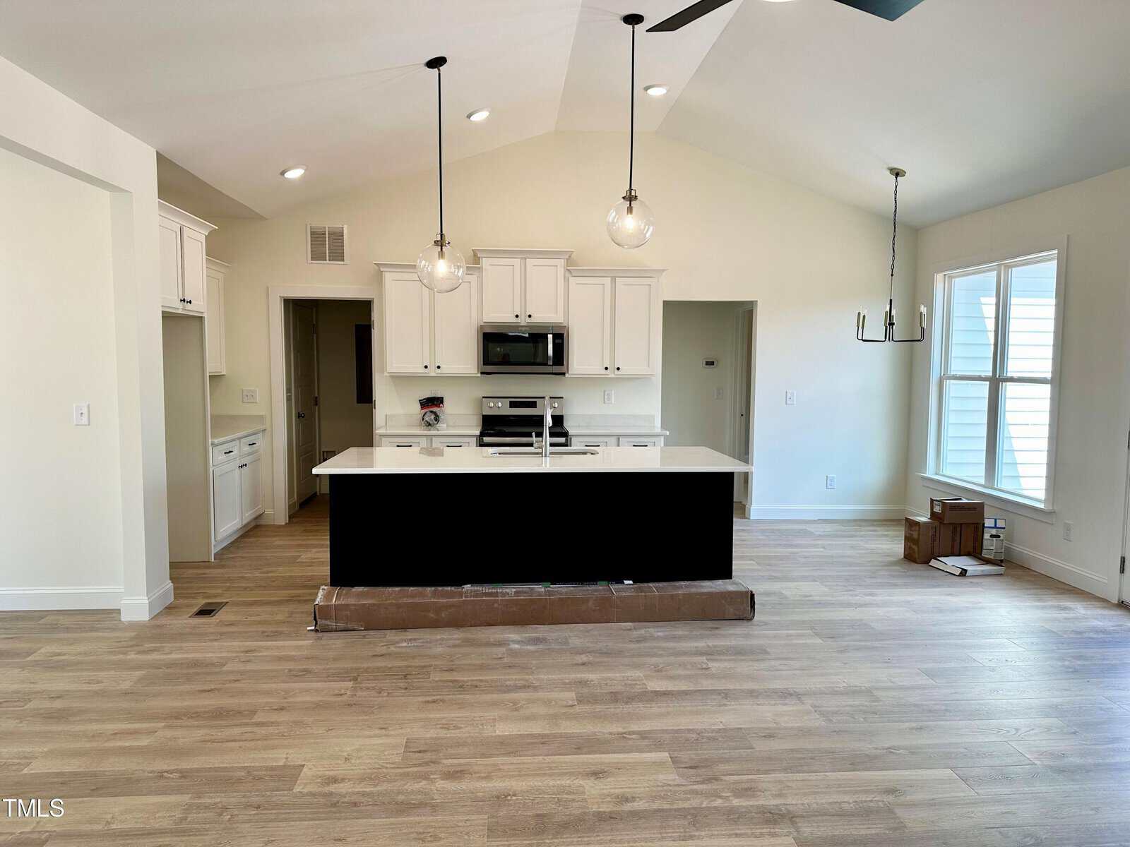 Modern kitchen with light wood floors, white cabinets, a black island with white countertop, pendant lights above, built-in appliances, and large windows letting in natural light. Cardboard boxes are stacked near the windows.