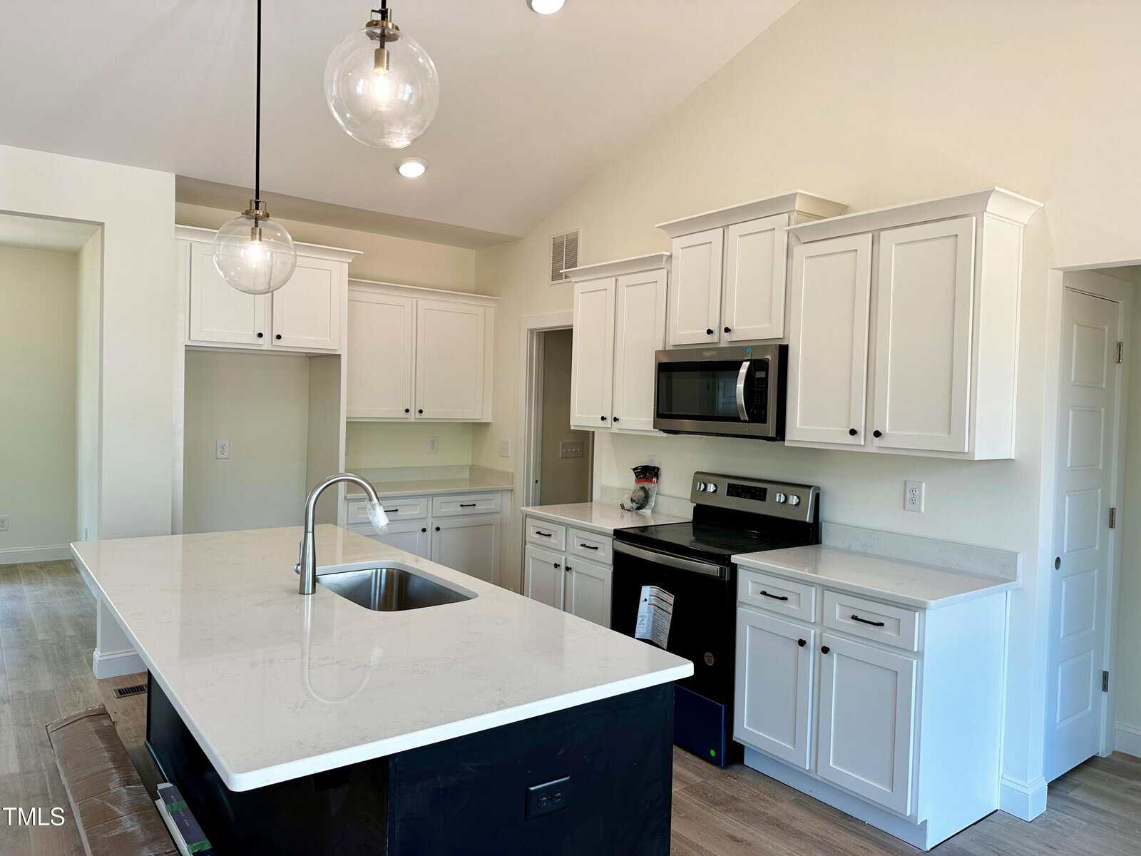 Modern kitchen with white cabinets, stainless steel appliances, a black island with a sink, pendant lights, and light-colored flooring. The counters are white, and the room has a bright, clean appearance.