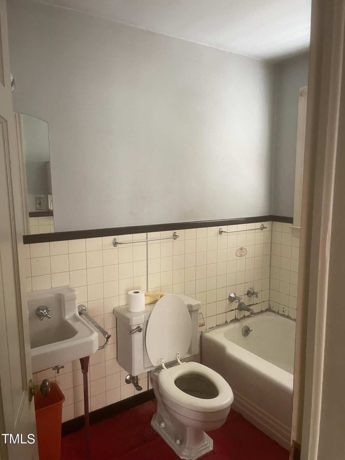 A small bathroom with white tiled walls and a red floor. Features a toilet with a toilet paper roll on top, a wall-mounted sink with faucet, a mirror, and a bathtub with a showerhead. The walls are light gray.