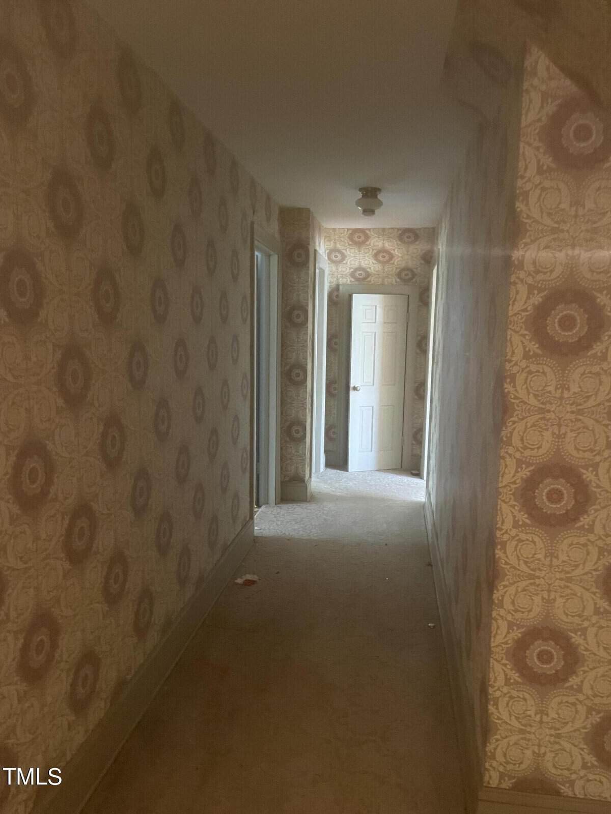 A dimly lit hallway with beige, patterned wallpaper and a carpeted floor. The corridor leads to a white door at the end, with additional doorways on the sides. A ceiling light fixture is visible.