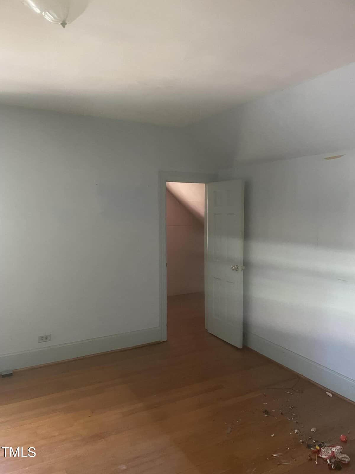 An empty room with light blue walls and a wooden floor. The room features a partially open white door leading to a small space or closet, and there is some debris on the floor near the bottom right corner. A light fixture is visible on the ceiling.