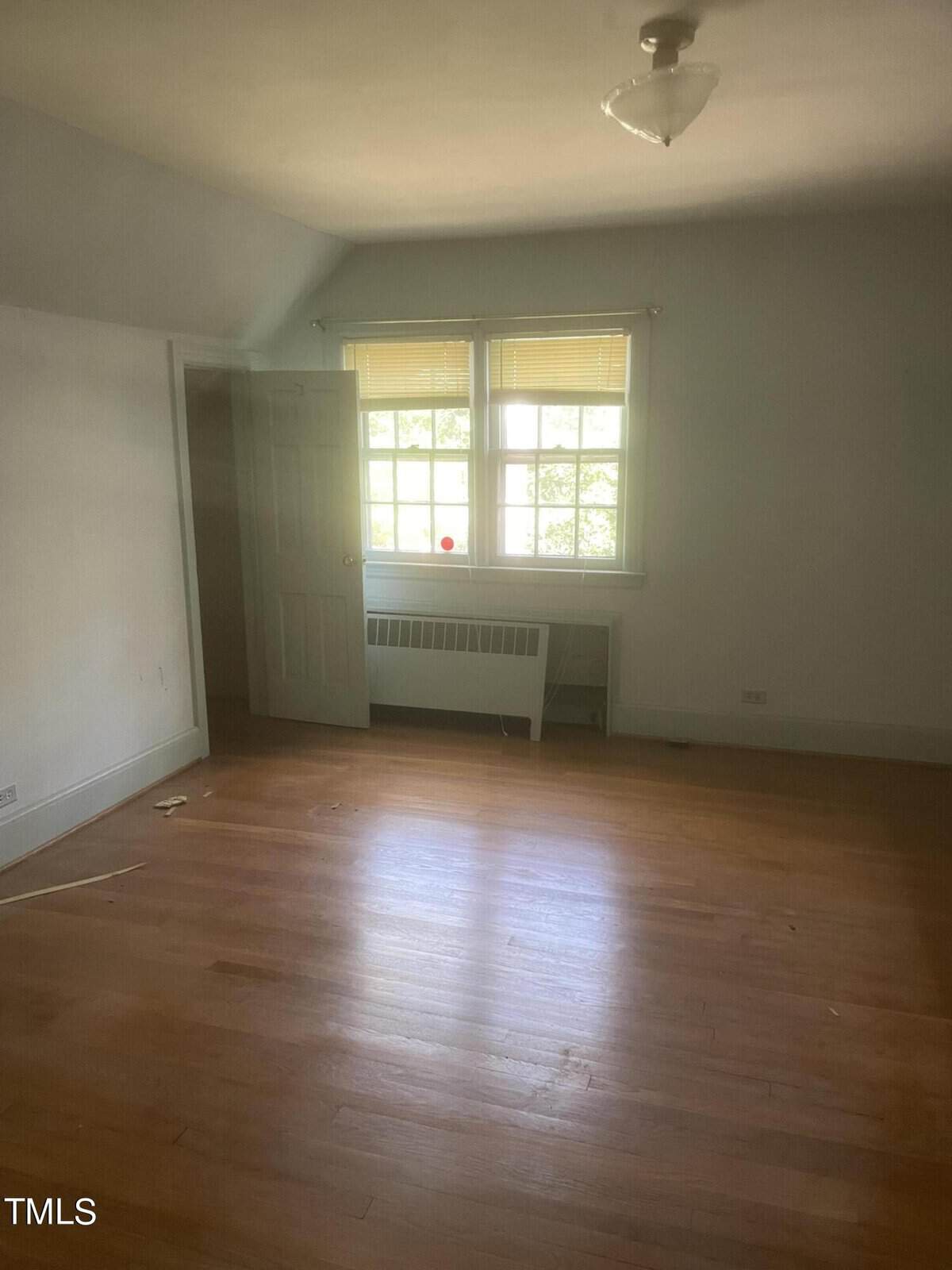 An empty room with wooden flooring and white walls. A door is slightly ajar on the left. A window with closed blinds is centered on the far wall, letting in light. A radiator is positioned below the window. Ceiling light fixture visible.