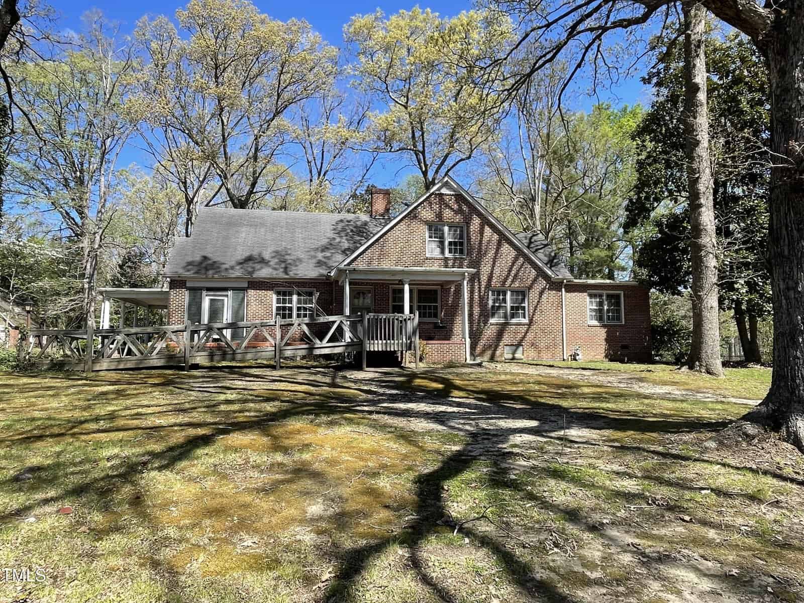 A brick house at 710 Hillboro Street, with a gabled roof, is surrounded by tall trees in sunny Oxford. The front yard features a wooden fence, casting shadows from the branches onto the ground. Above, the sky is clear and blue.