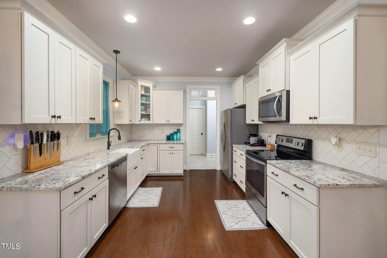 Modern kitchen with white cabinets, granite countertops, and stainless steel appliances. A knife block sits on the left counter, and a teal dish towel hangs by the window. Hardwood floors and recessed lighting complete the bright, spacious design.