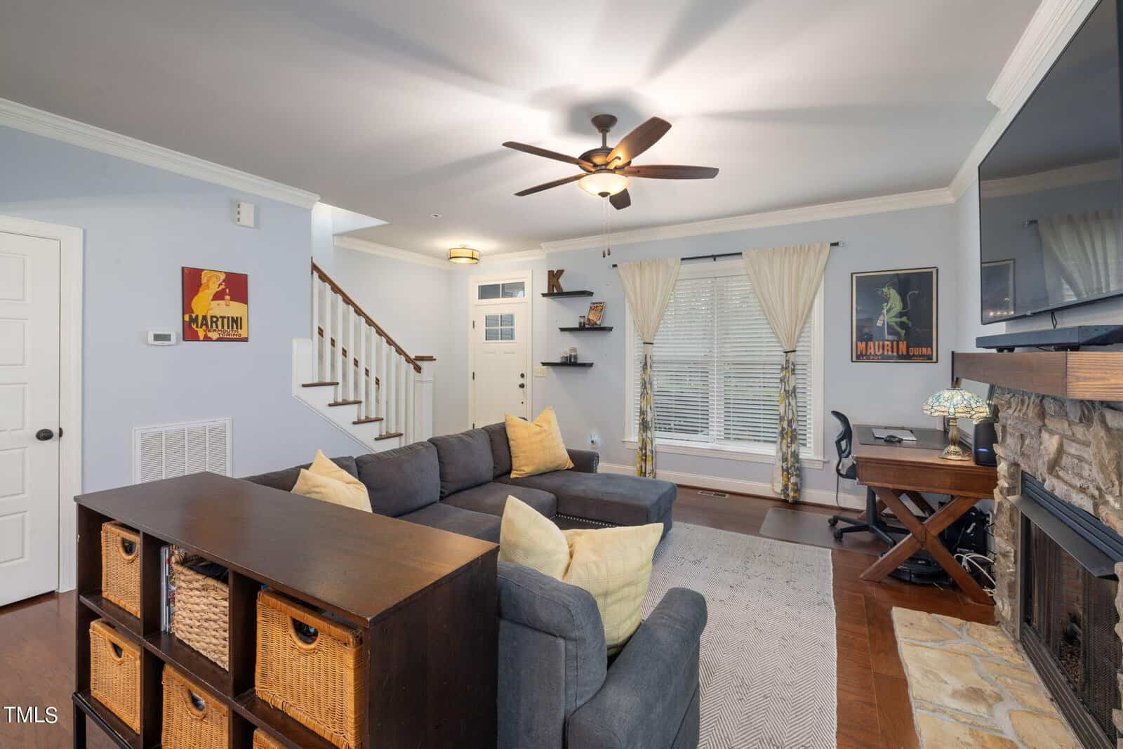 A cozy living room features a sectional sofa with cushions, a wooden storage unit, a stone fireplace, and a TV. A small desk with a chair sits near a window with sheer curtains. Stairs lead up, and posters adorn the walls.