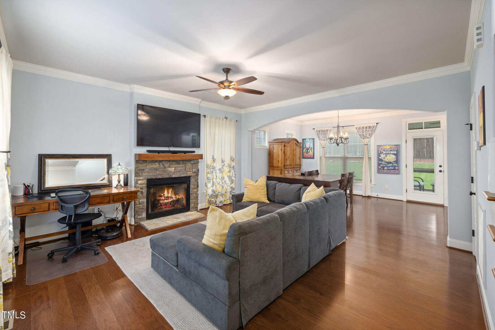 A cozy living room with a gray sectional sofa and yellow throw pillows faces a stone fireplace with a TV above it. A small wooden desk with a chair is on the left. The room opens to a dining area with a chandelier and large windows.